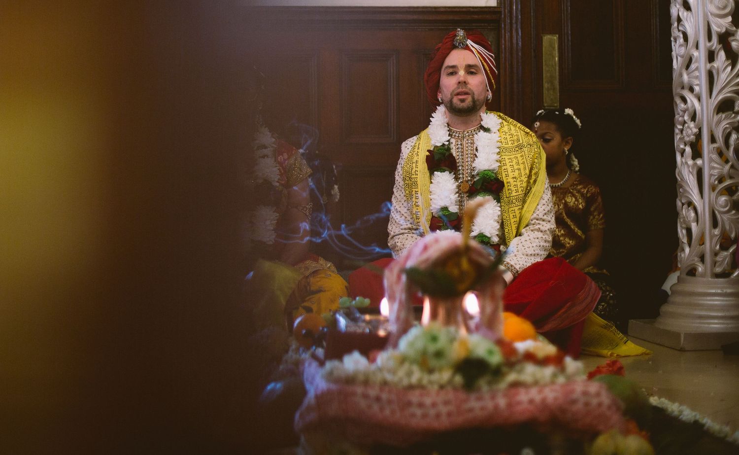 A figure in bright yellow and red attire sits in meditation with ornate decorative elements in a dimly lit setting.