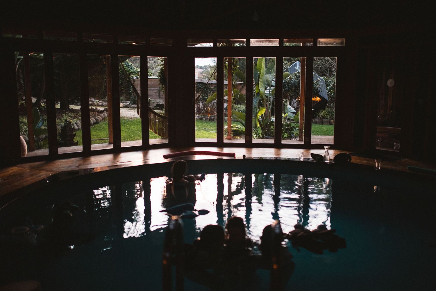 Wide panoramic view of an indoor swimming pool with dark silhouettes against bright windows.