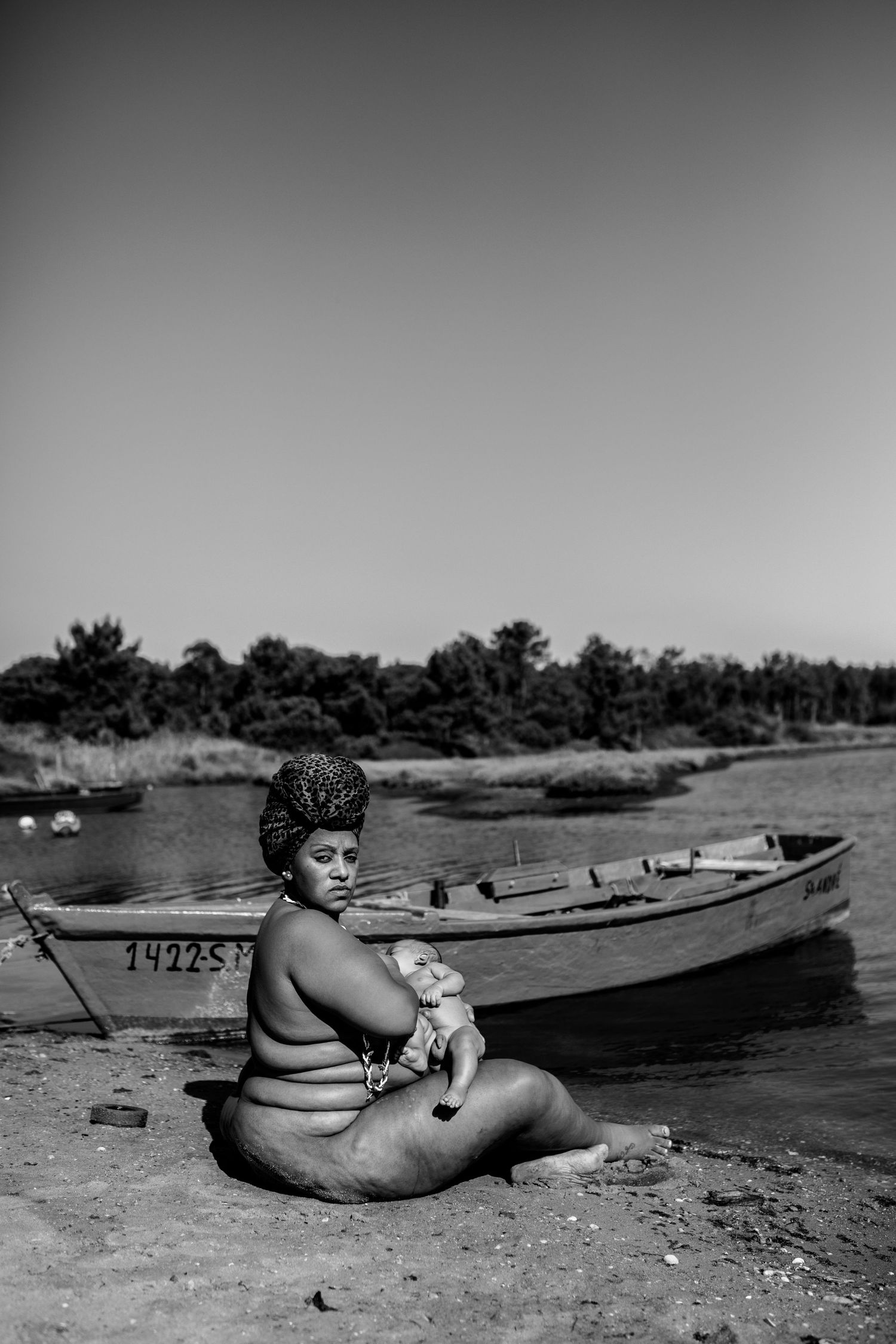 A vintage black and white photograph of someone sitting by a lake shore with boats in the background.