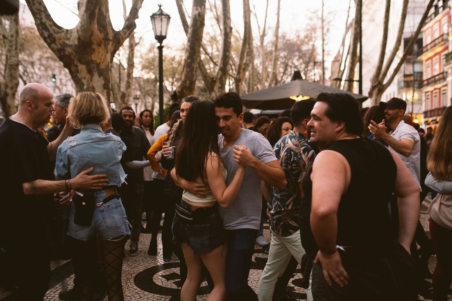 A lively crowd gathers outdoors beneath trees near a street lamp during a social event.