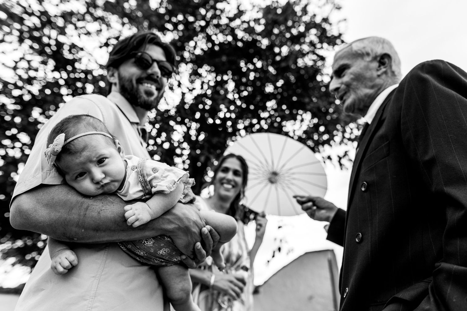 A candid black and white photo of a tender family moment outdoors under trees.