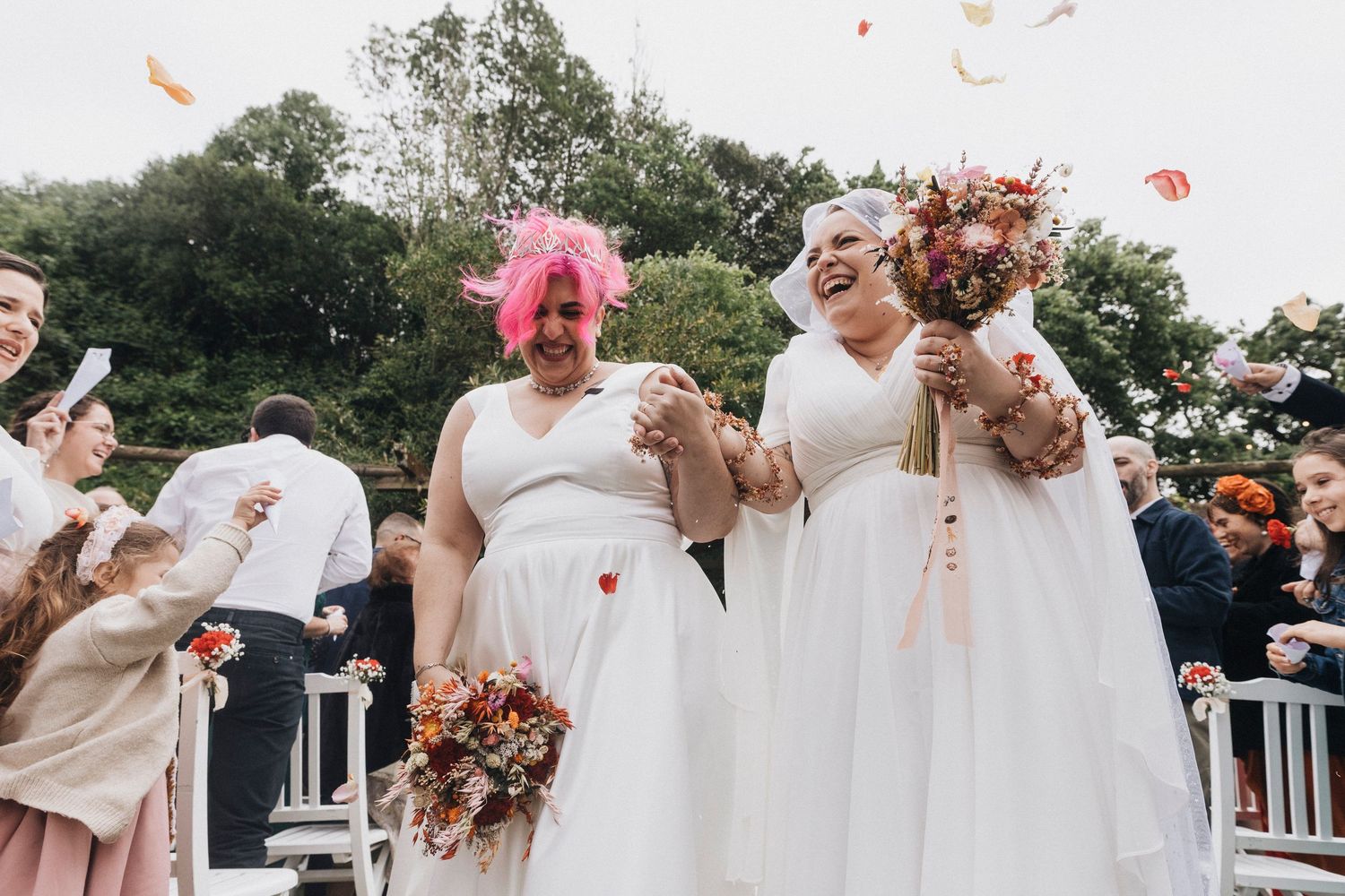 Wedding party celebrating with flower petals and confetti in white outfits against cloudy sky.