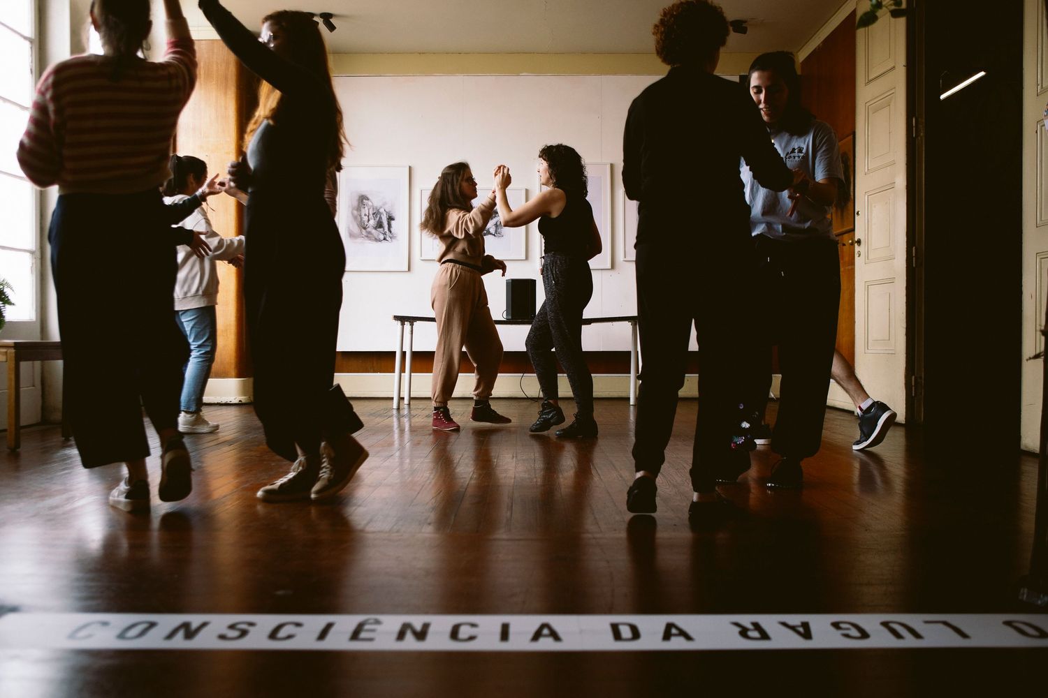 People dance in a dimly lit studio space with silhouettes visible against bright windows and reflective floor.