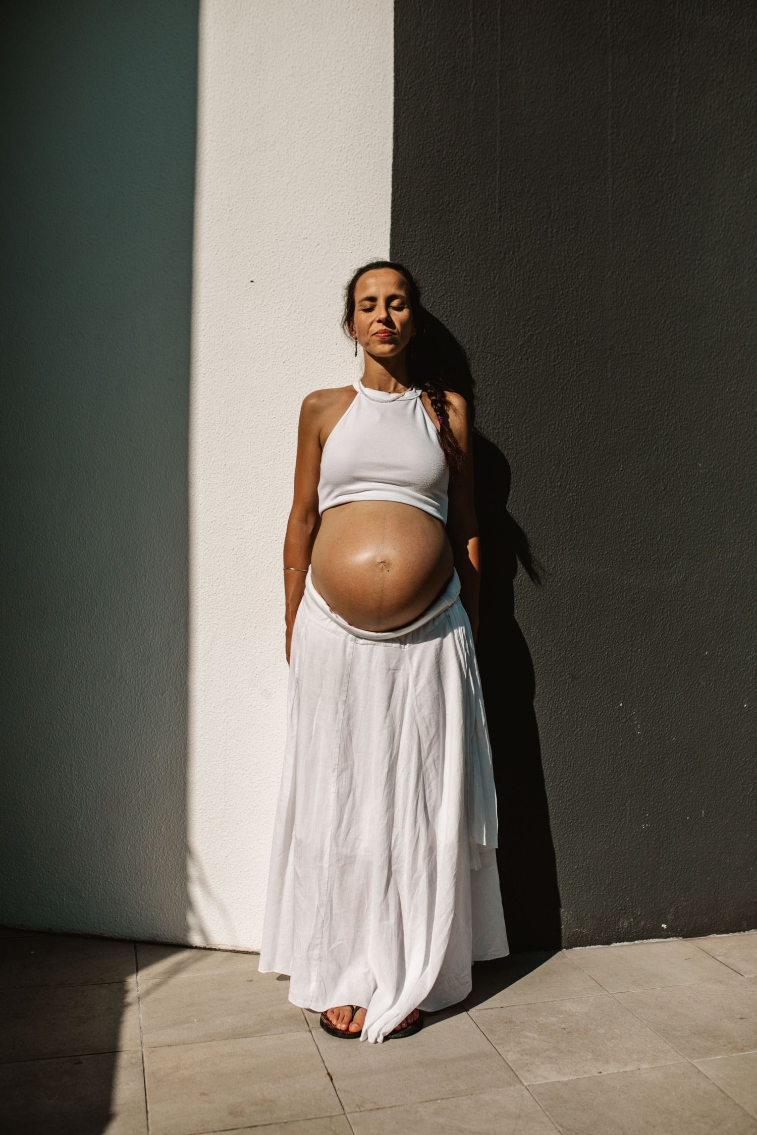 Minimalist maternity photography against a stark wall with dramatic shadows and white clothing.