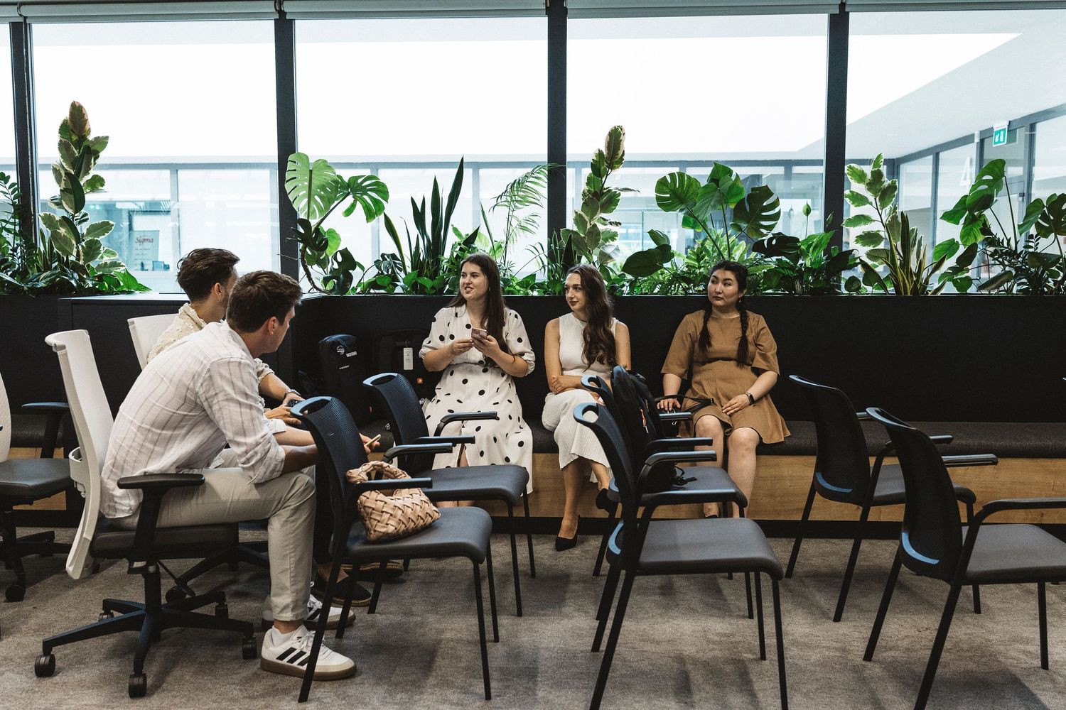 Modern office space with people working and collaborating at desks surrounded by indoor plants and natural lighting.