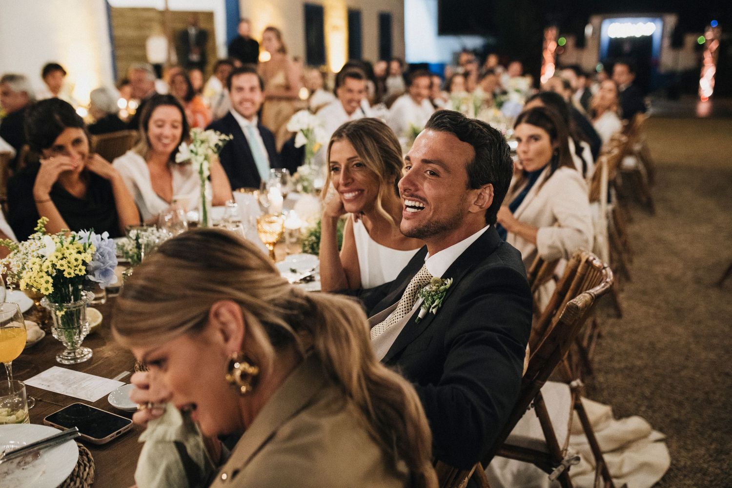 Wedding reception guests enjoy dinner at long tables under warm lighting.
