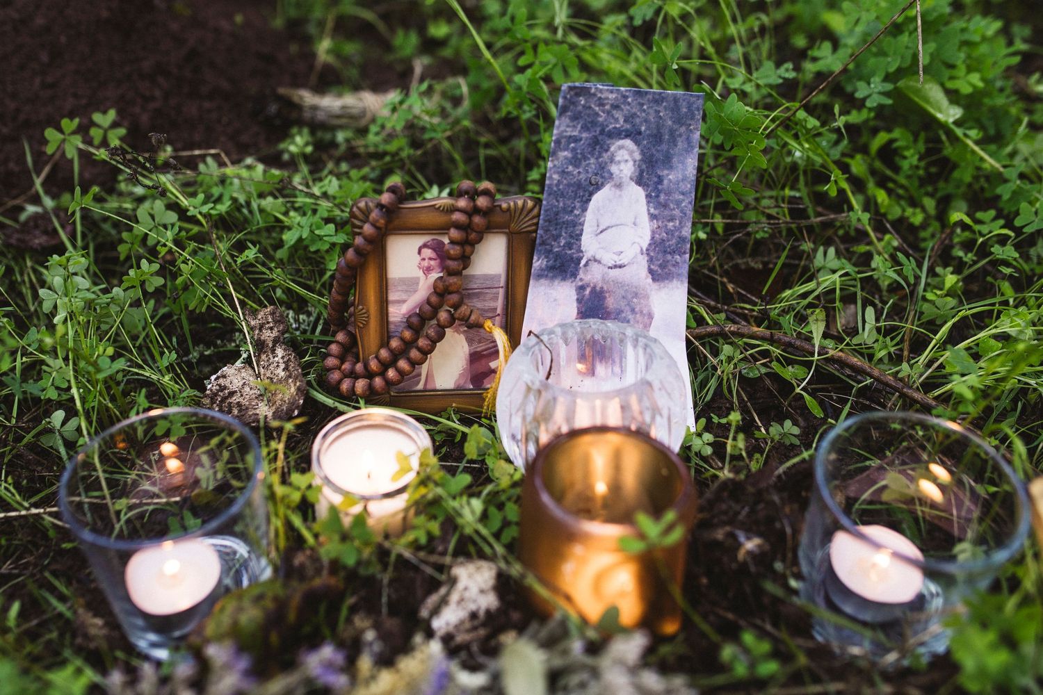Memorial display with candles, photographs and personal items arranged on mossy ground in a garden setting.