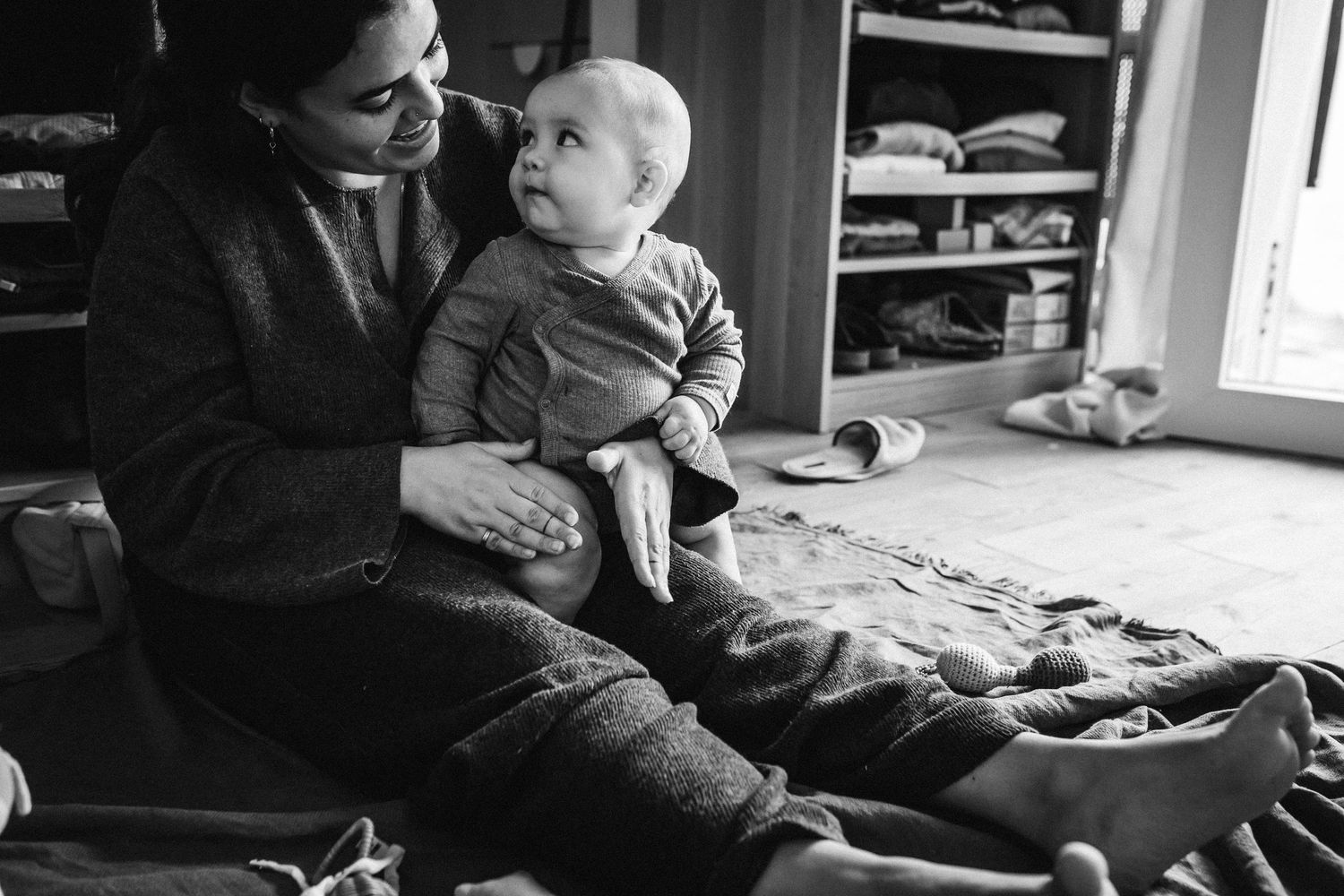 A black and white photograph shows a tender moment between an adult and baby sitting together on the floor.