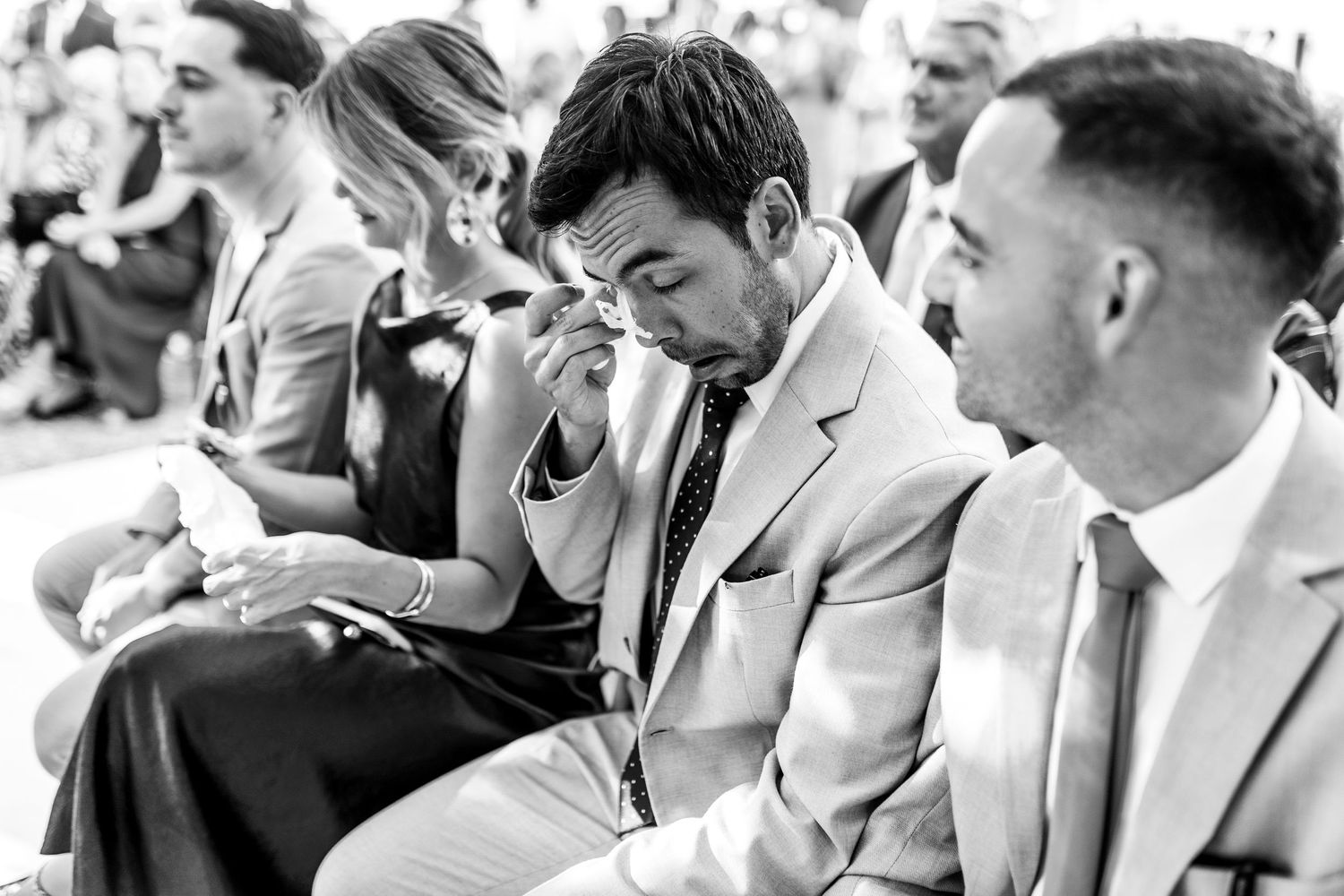 Wedding guests in formal attire gather outdoors for a celebration on a sunny day.
