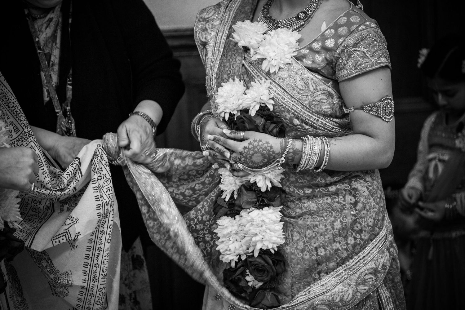 Black and white photo showing traditional Indian wedding attire with detailed henna designs and white floral garlands.
