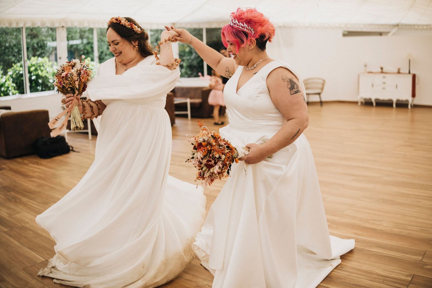 Two people in flowing white dresses dance joyfully together in a sunlit room with wooden floors.