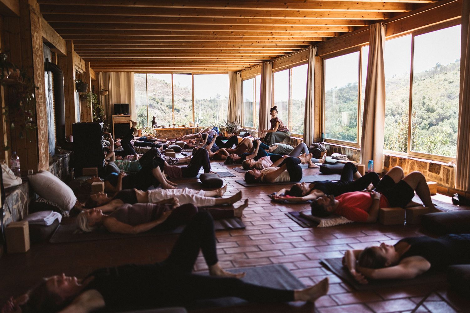 Group of people practicing yoga and meditation in a sunlit wooden studio with large windows overlooking nature.