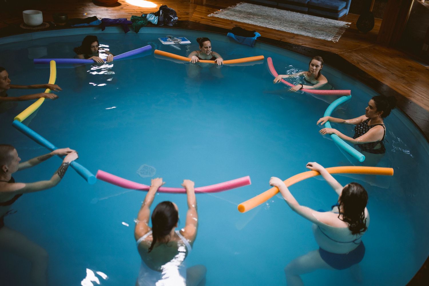 People floating and relaxing in bright blue pool water with colorful pool noodles.