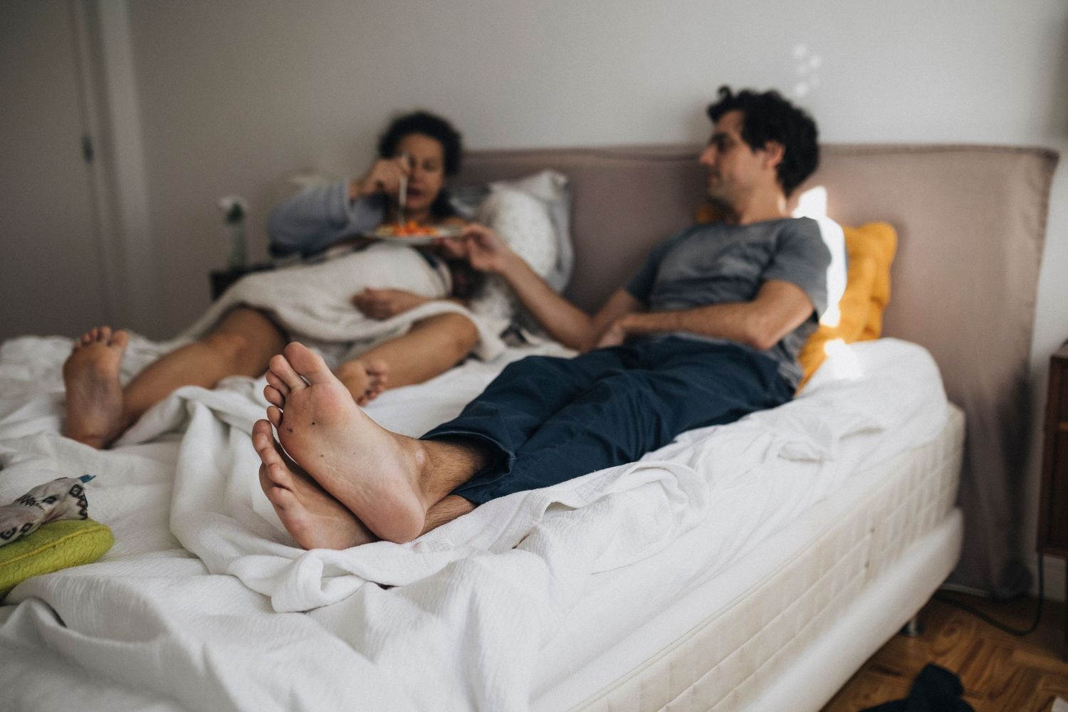 Person sleeping in bed with sunlight streaming through window while wearing white t-shirt and dark pants.