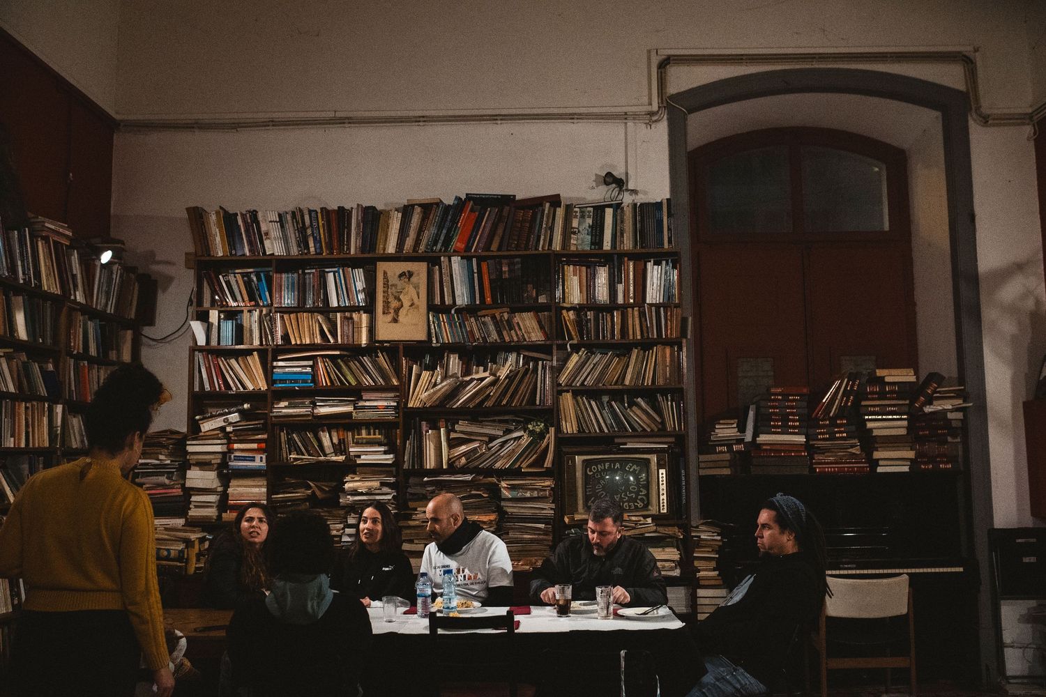 A dimly lit library or bookstore interior with rows of bookshelves and people seated at tables.