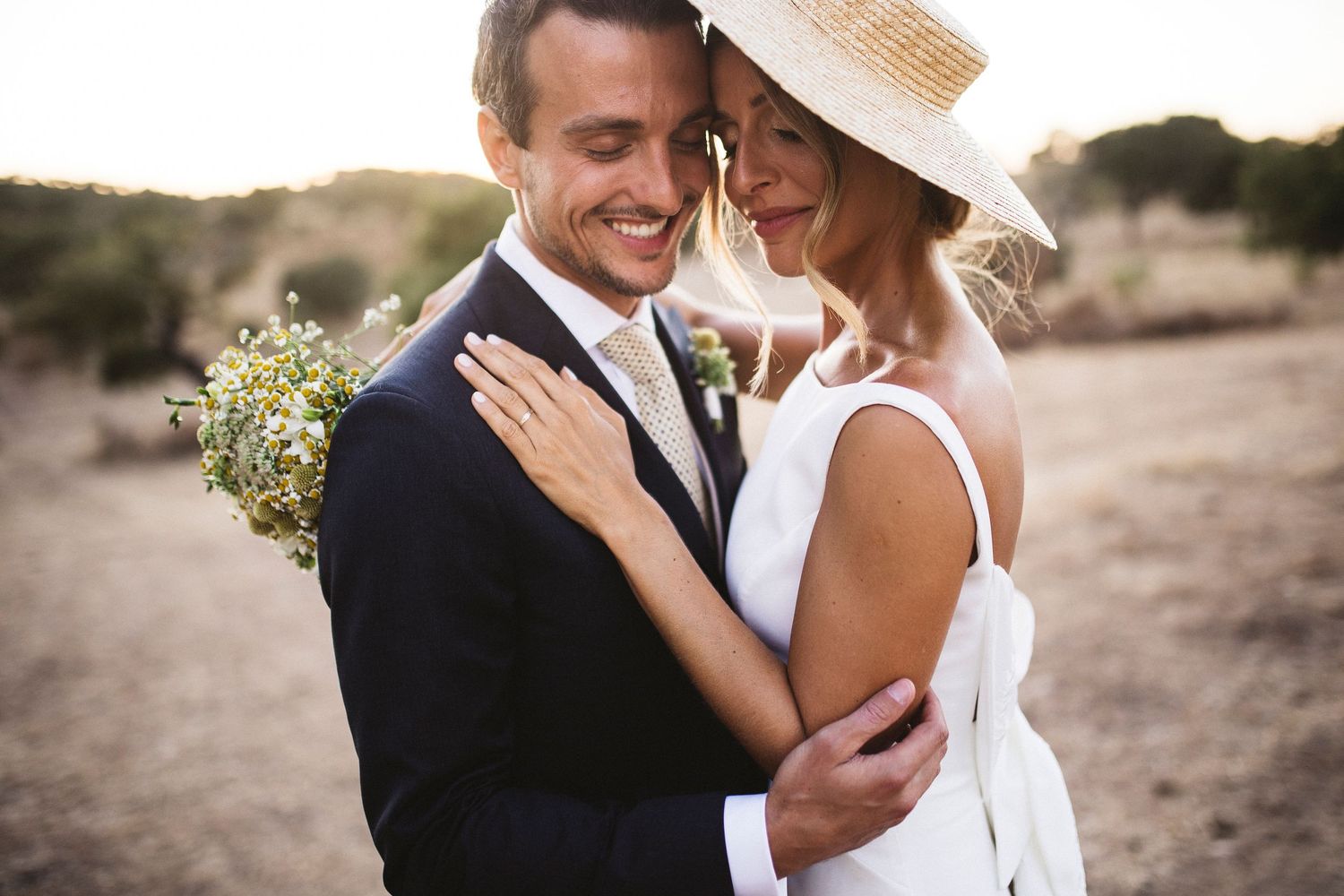 Bride in white hat and groom share joyful moments during outdoor portraits.