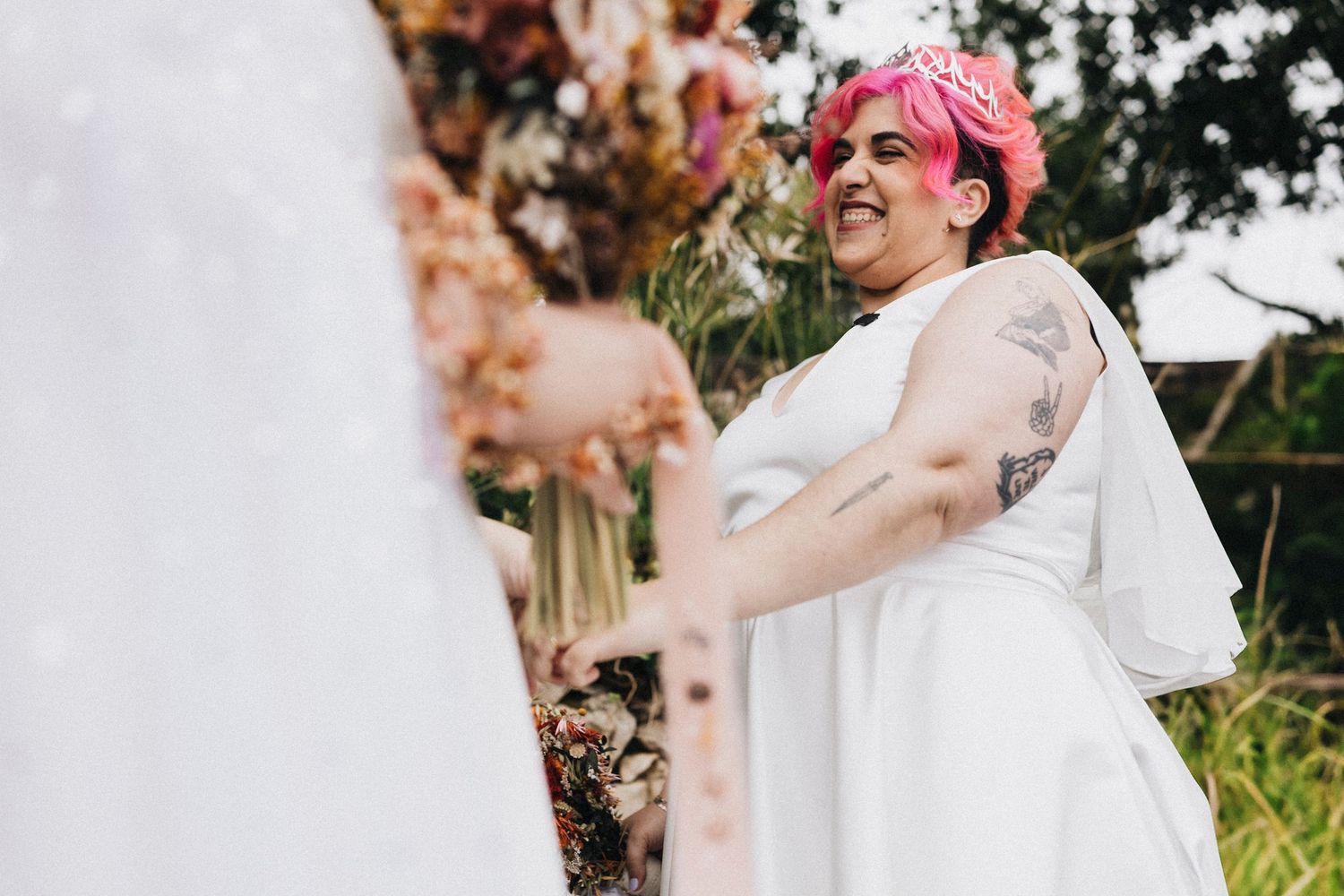 Wedding photo of person in white dress with pink hair accessory smiling outdoors.