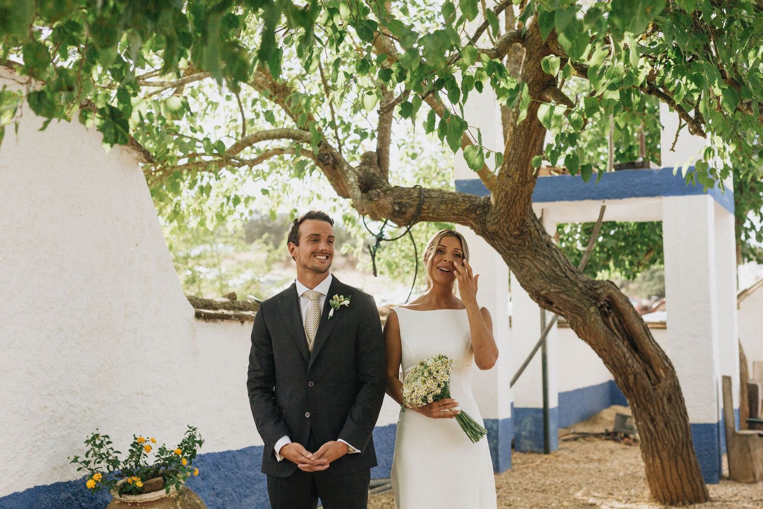 A couple poses under a tree during their outdoor wedding at a rustic white venue.