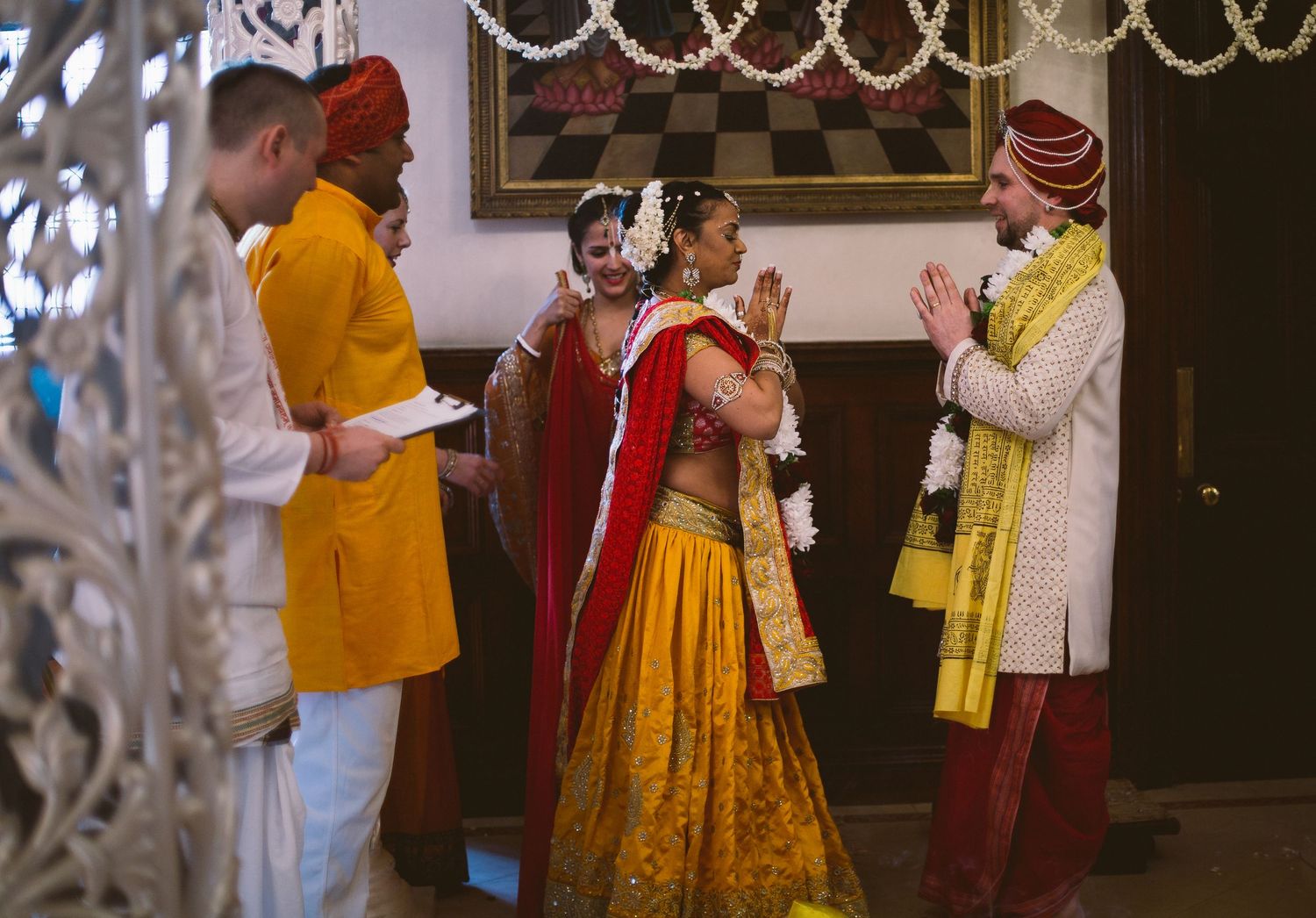 Traditional Indian wedding ceremony participants in colorful attire gather indoors near ornate decorative elements.