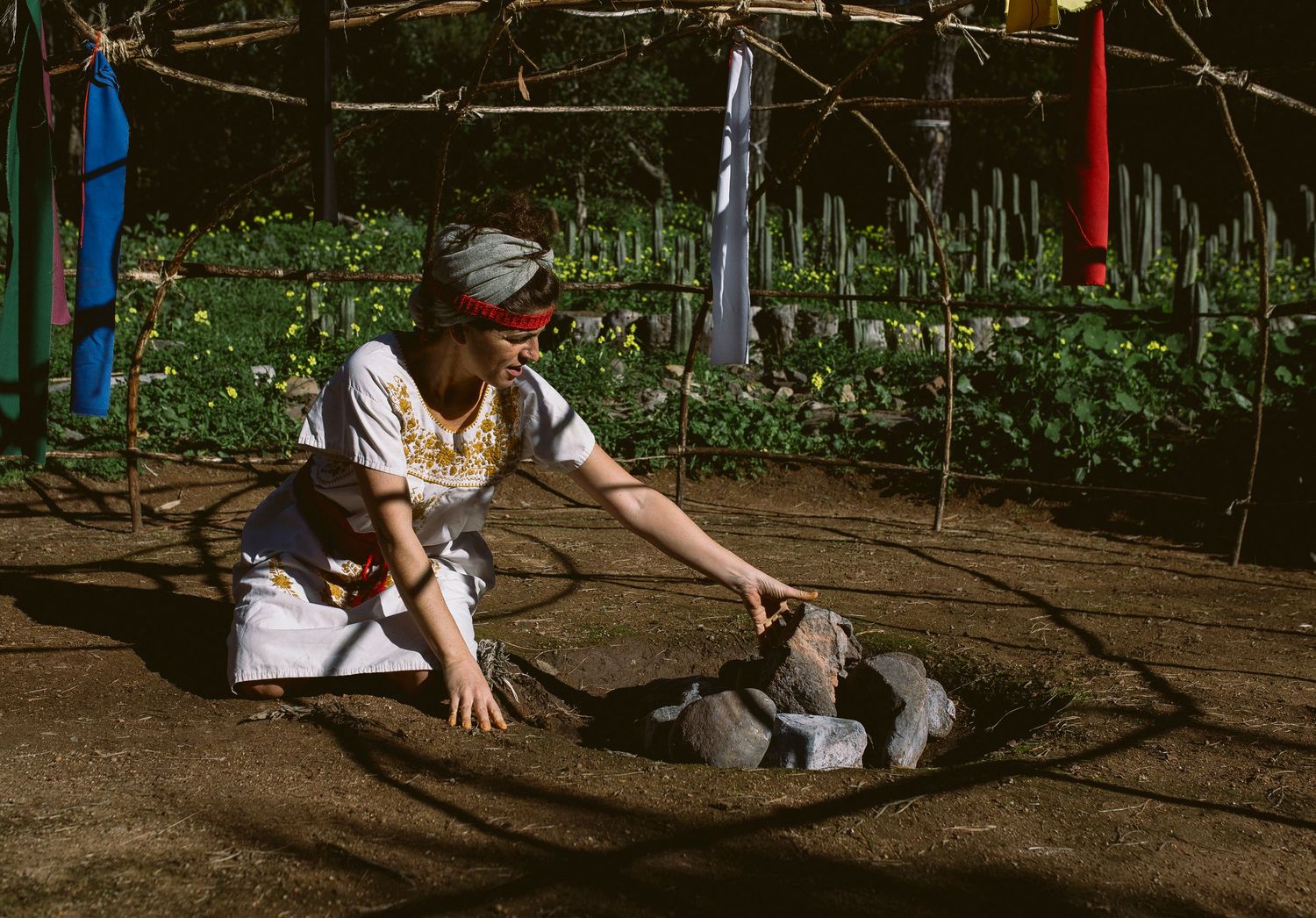 A person sits on the ground in a garden area surrounded by wooden raised planting beds and gardening tools.