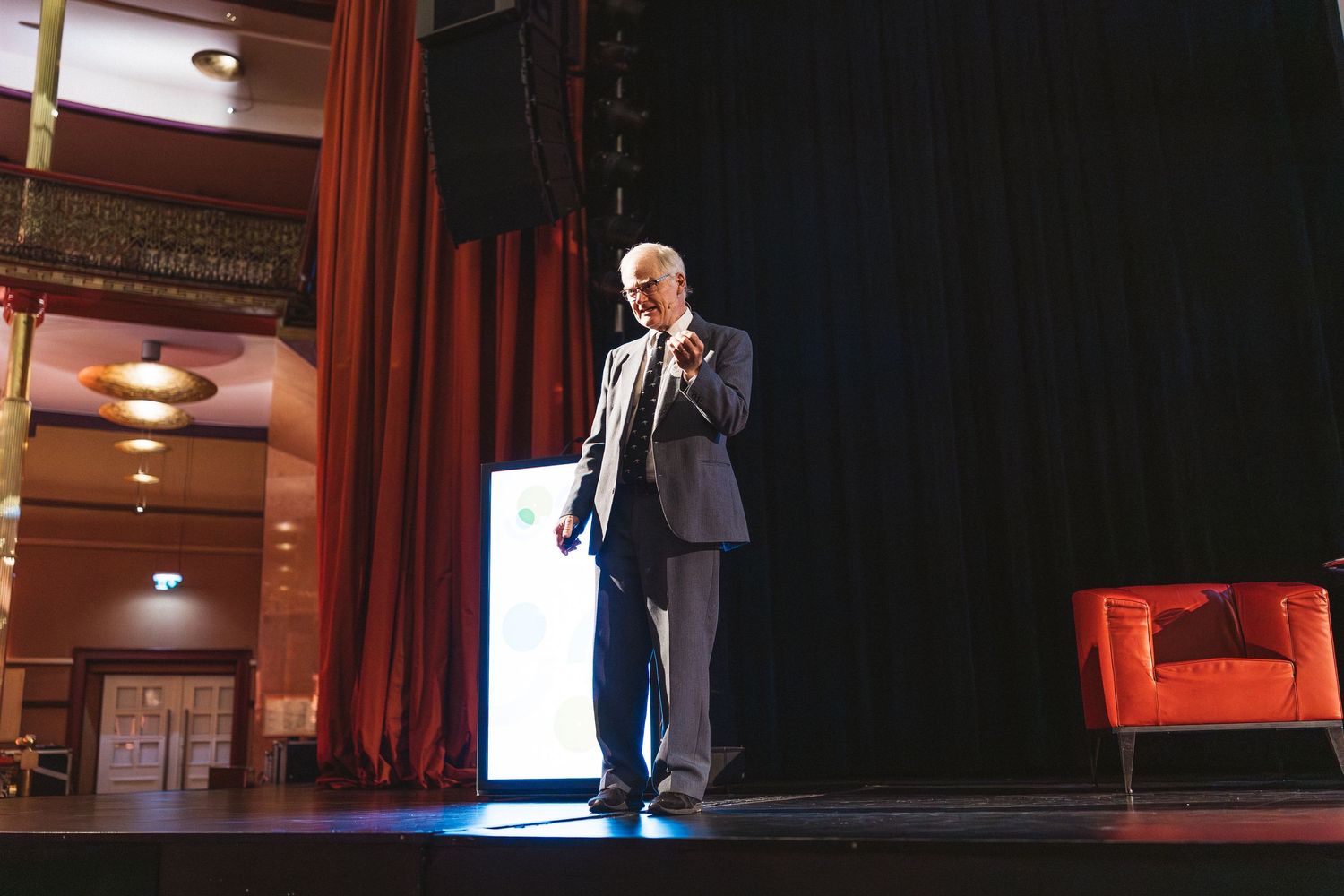 A person gestures while giving a presentation on stage in a dark auditorium with red curtains and screens.