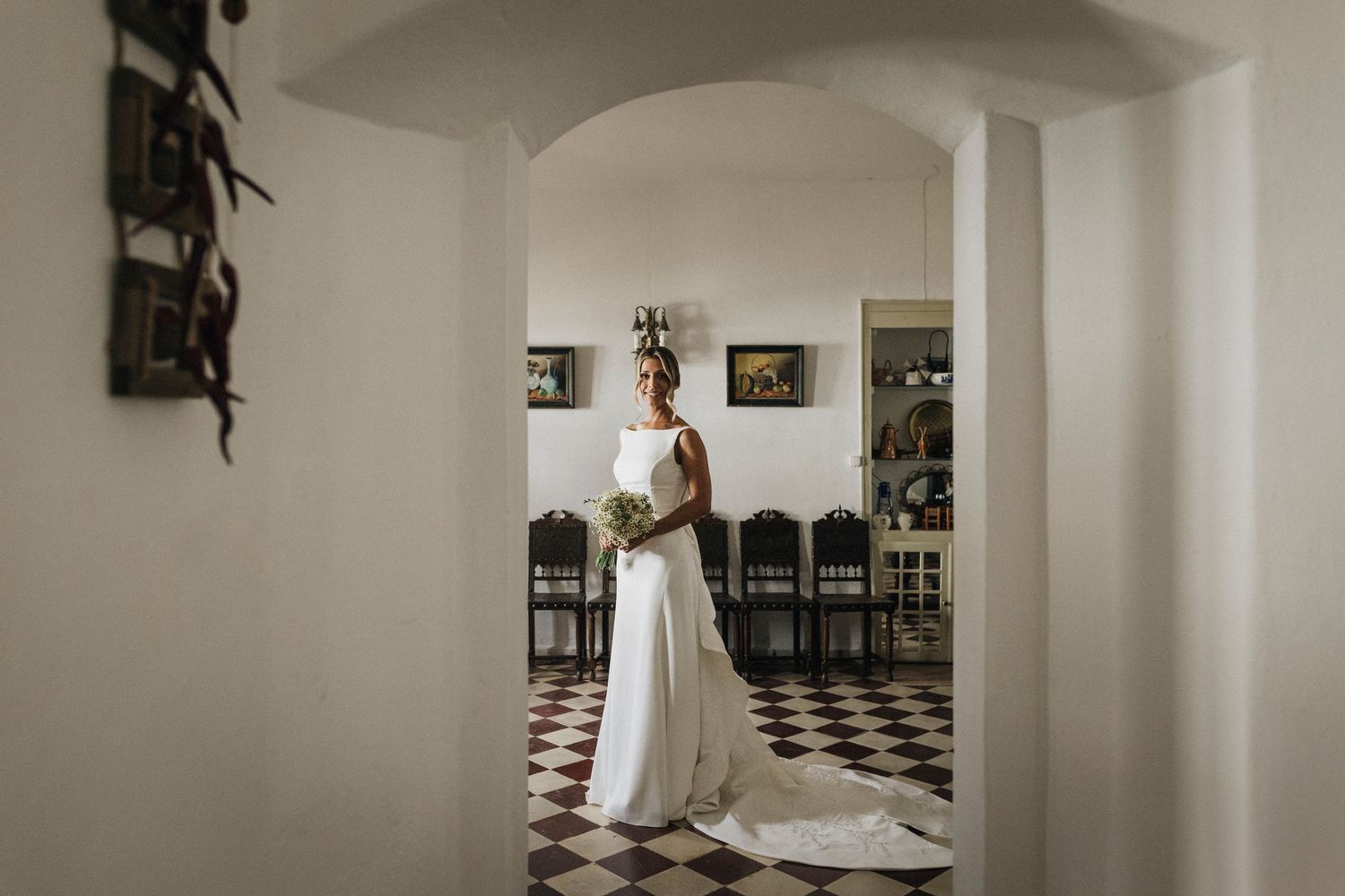 Elegant wedding dress silhouette framed in a doorway with black and white checkered floor and arched ceiling.