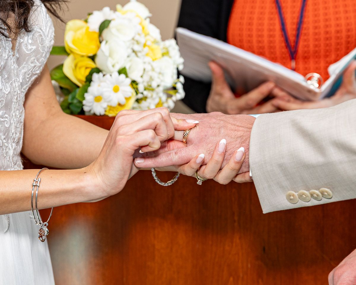 Close-up of hands exchanging wedding rings during ceremony with yellow and white bouquet in background.