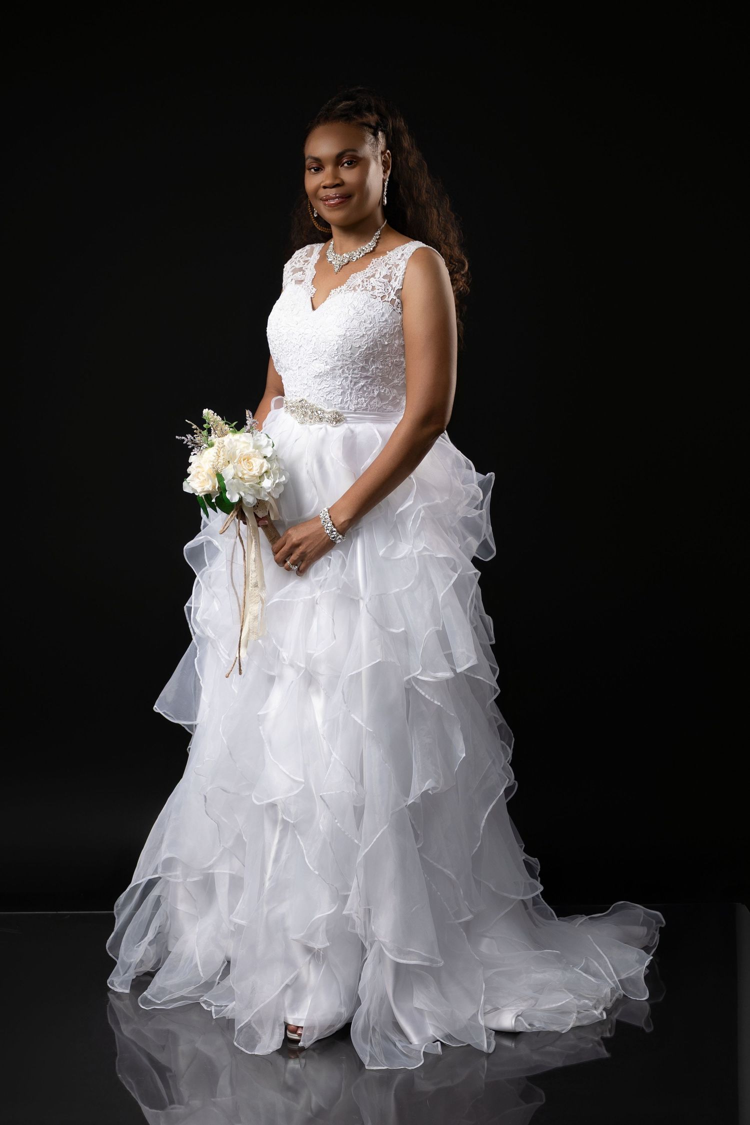 A bride in a flowing white ruffled wedding gown poses elegantly against a dark background in a bridal portrait session.