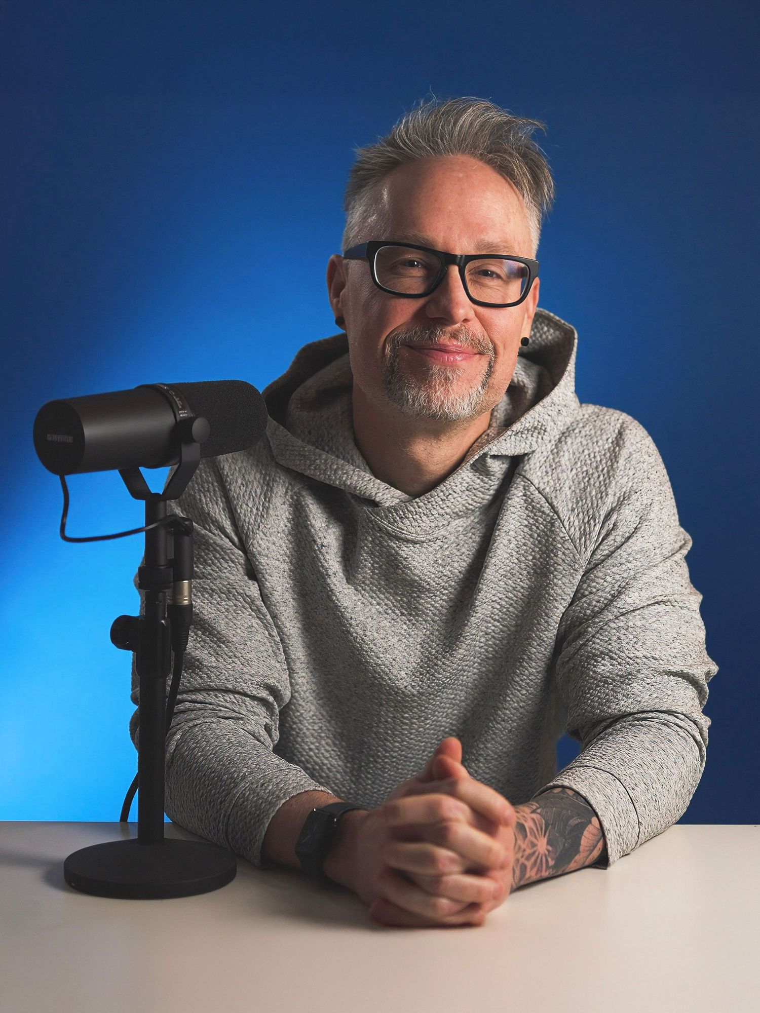 Middle-aged man with glasses and tattoos sits at a white desk with a podcast microphone against a blue background.