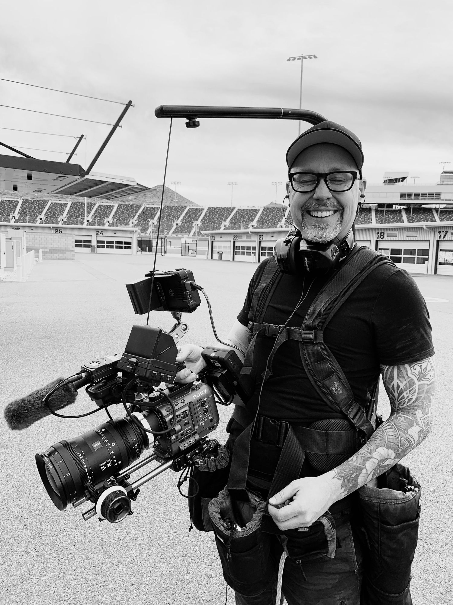 Tattooed cameraman smiling outdoors at a racetrack, holding a professional film camera with accessories, wearing a black cap.