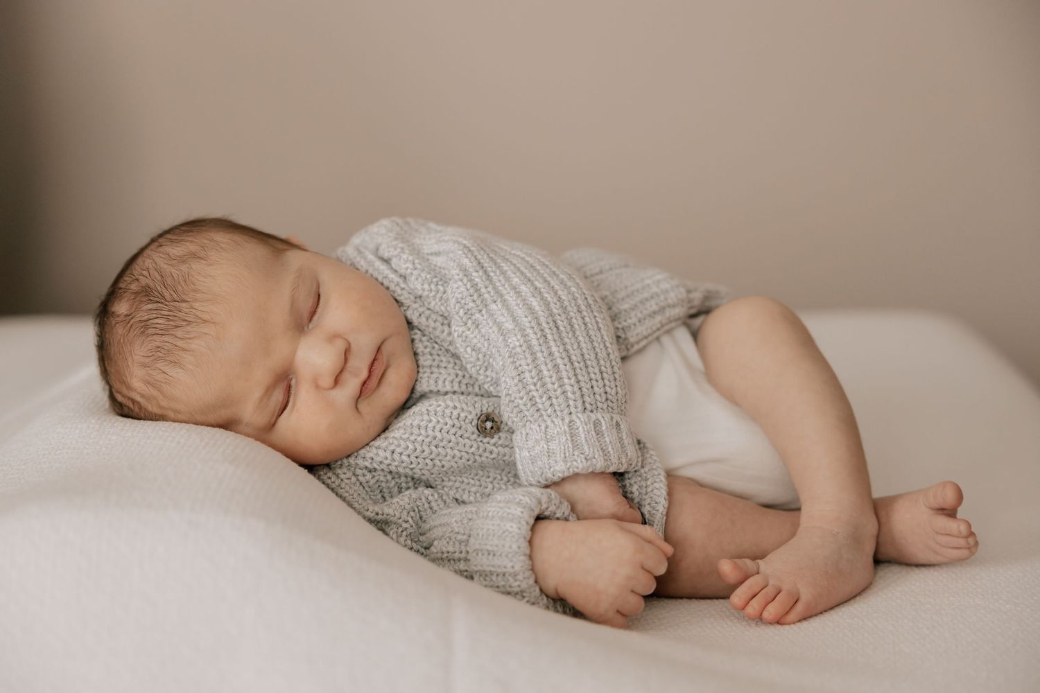 Peaceful sleeping newborn baby wearing a soft gray cardigan sweater rests on white bedding in natural light.