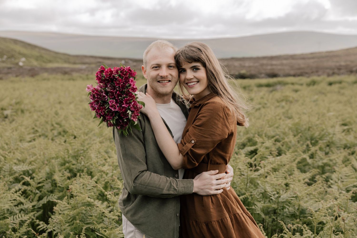 Two people embrace in a rustic field while holding vibrant red flowers against a moody countryside backdrop.