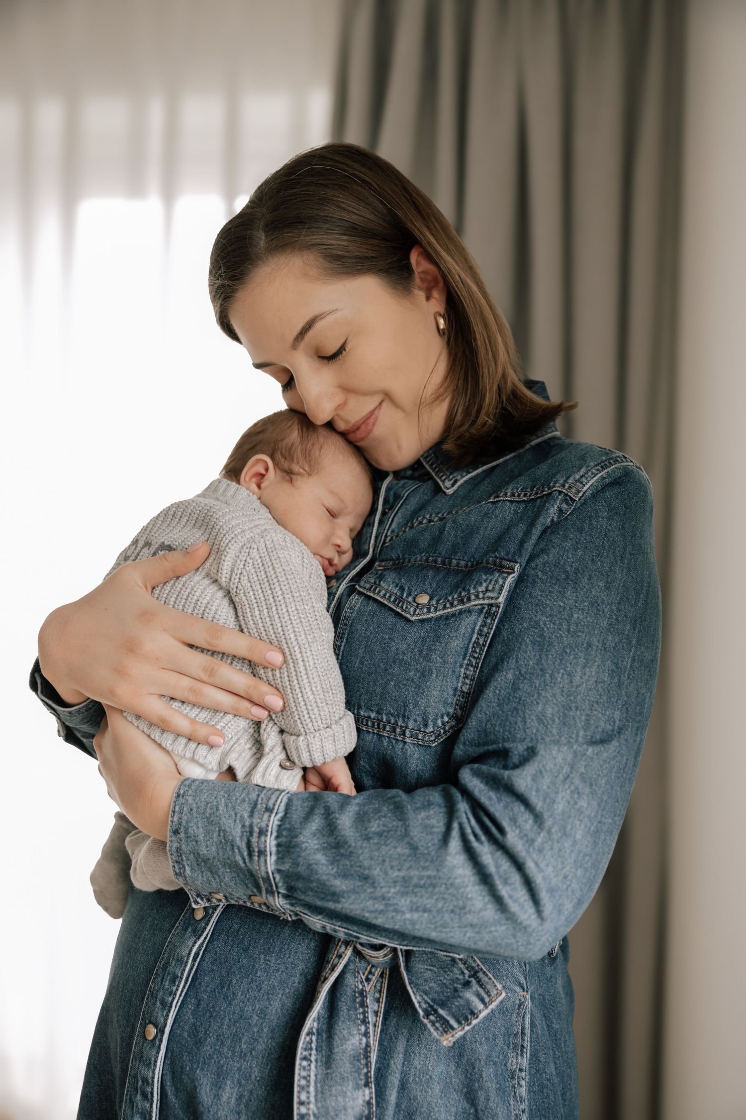 Person wearing denim jacket tenderly embraces and snuggles with sleeping baby in front of curtained window.