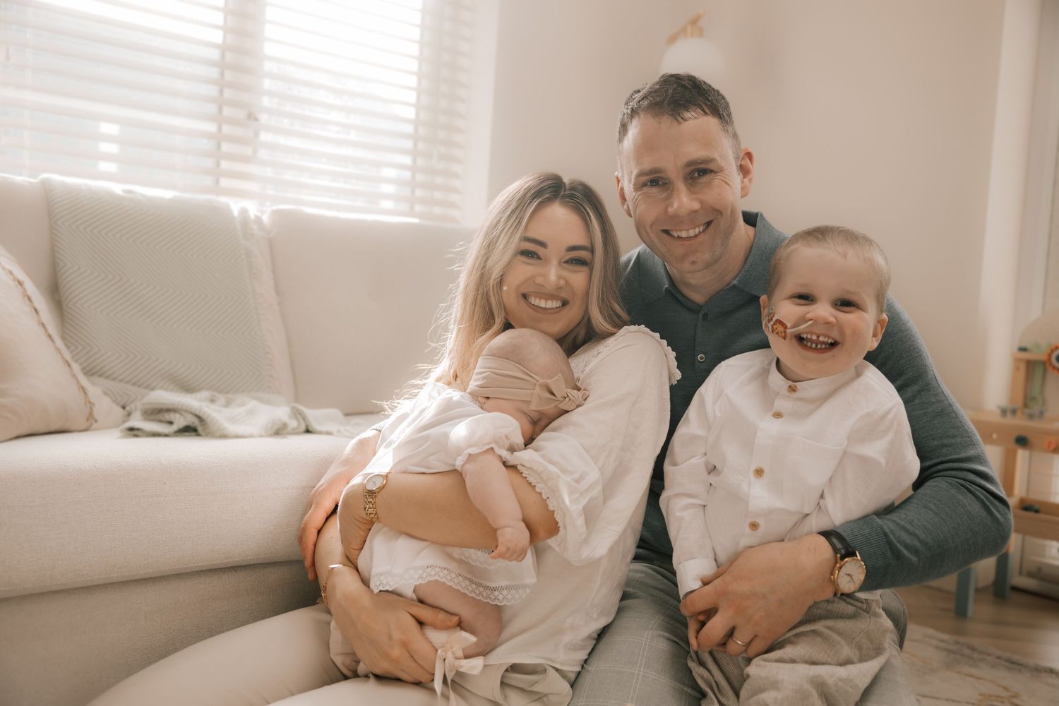A family lounges together on a couch in a bright, cozy living room with natural lighting through venetian blinds.