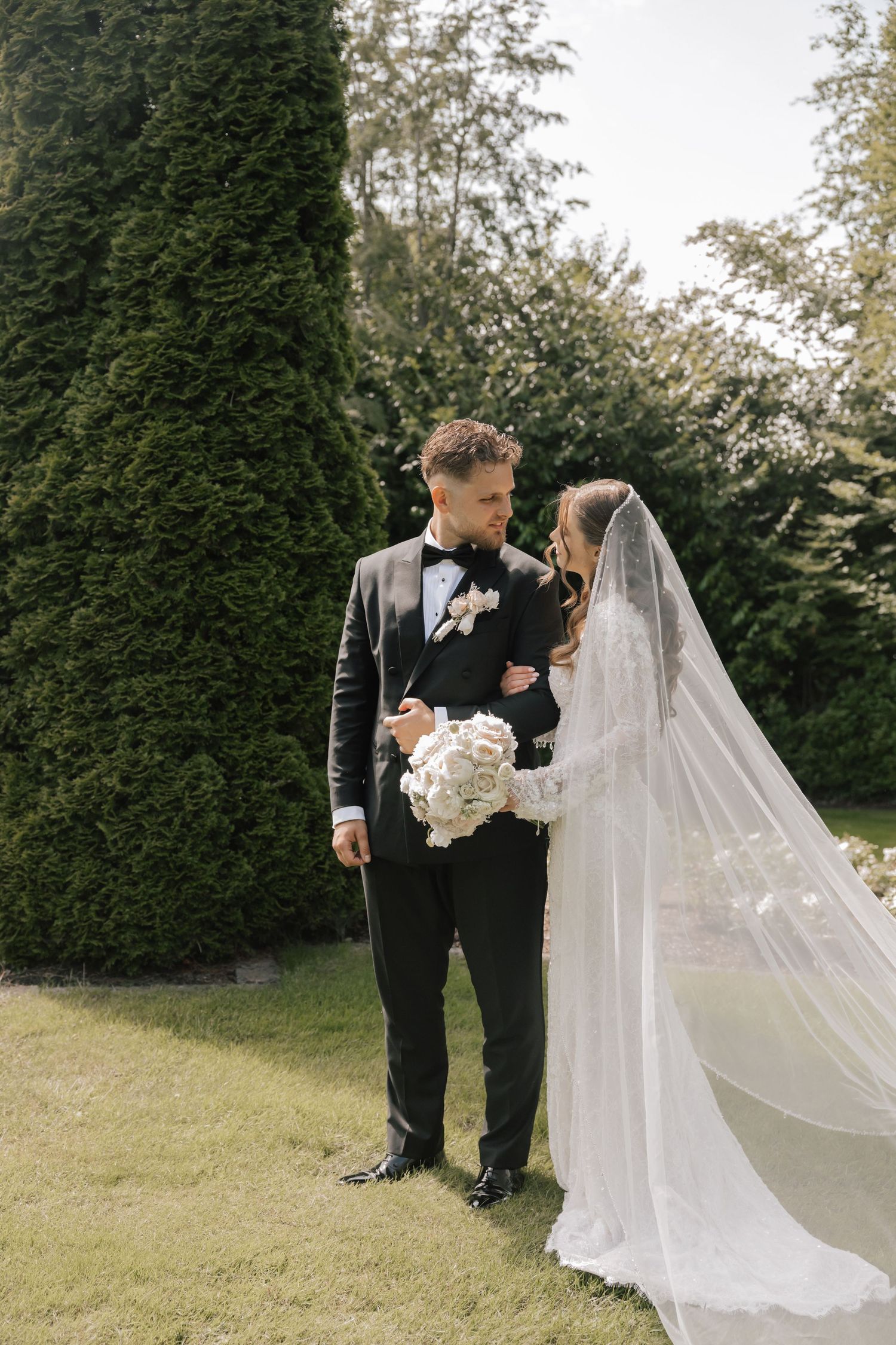 Bride and groom standing together in a lush garden, bride wearing a flowing veil and holding a white bouquet.