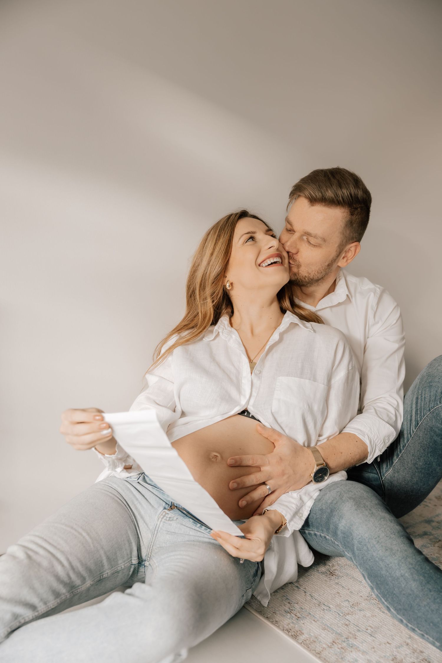 A joyful couple wearing white shirts embrace intimately while relaxing together on a light colored surface.
