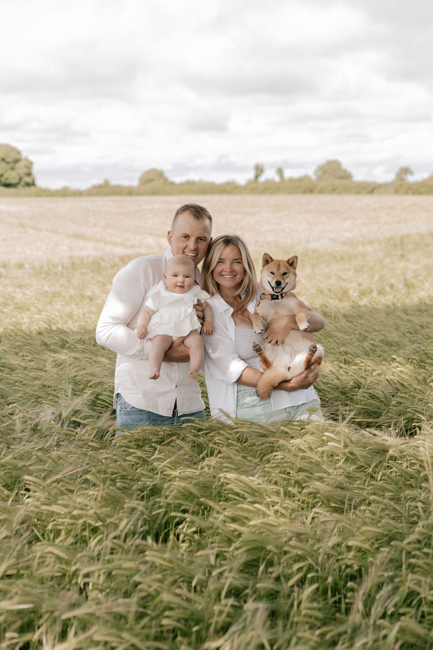 A family poses together with their dog in a golden wheat field on an overcast day.