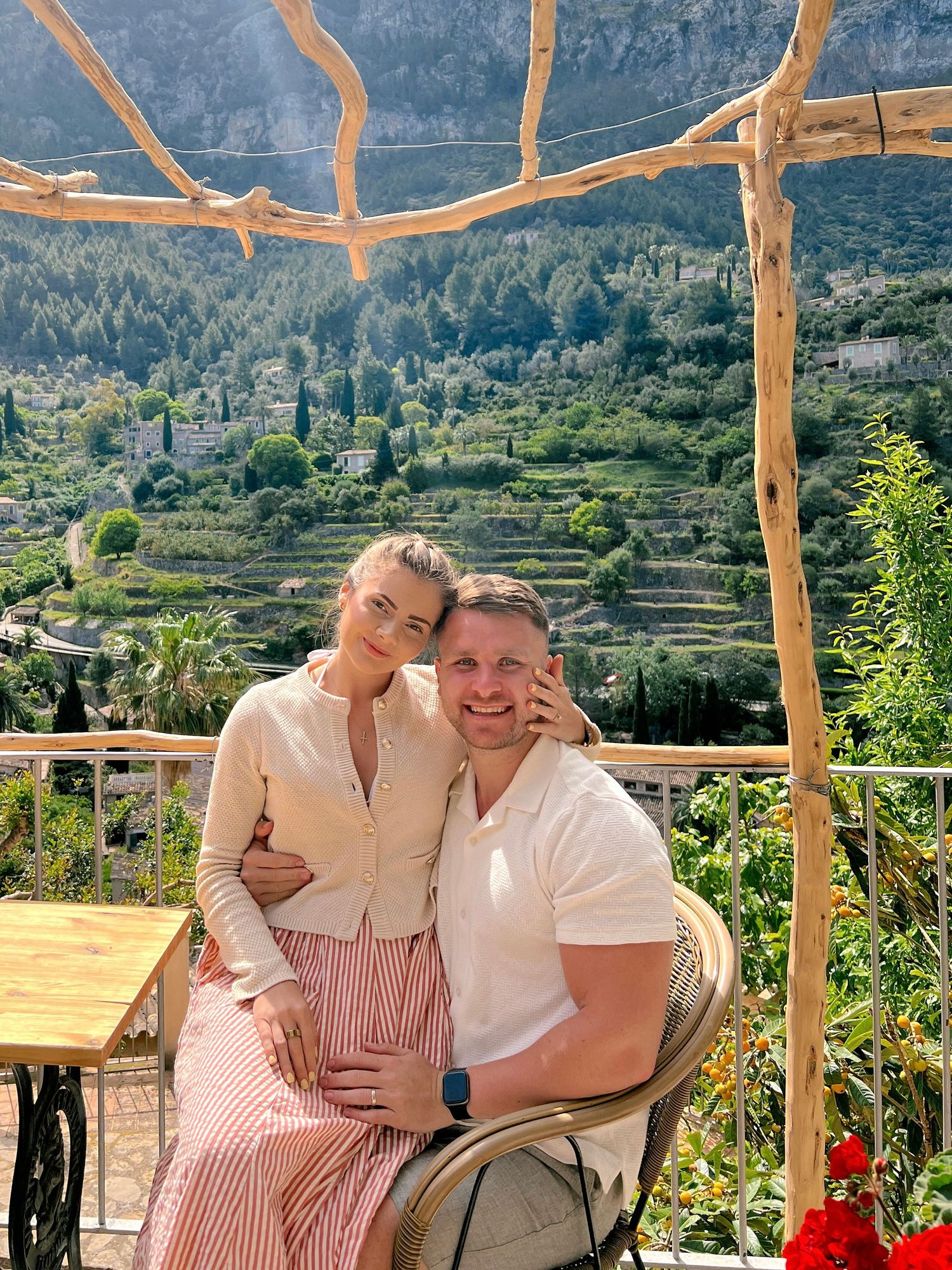 A couple enjoys a scenic view of terraced hillside gardens from a rustic wooden pergola on a sunny day in the Mediterranean.