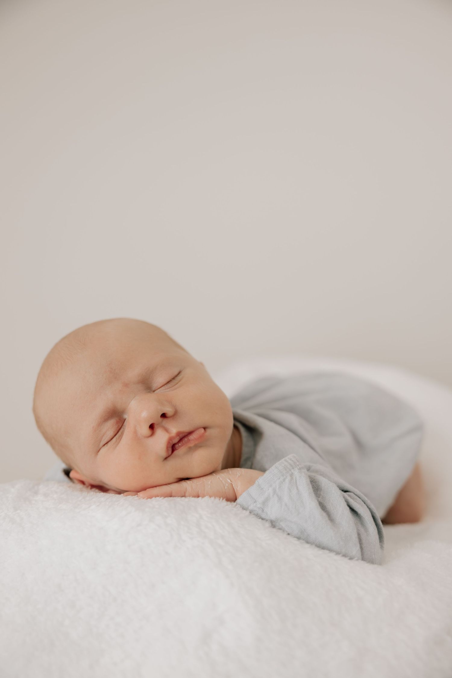 Peaceful newborn baby sleeping on a white fluffy blanket wearing a soft gray onesie in a serene portrait.