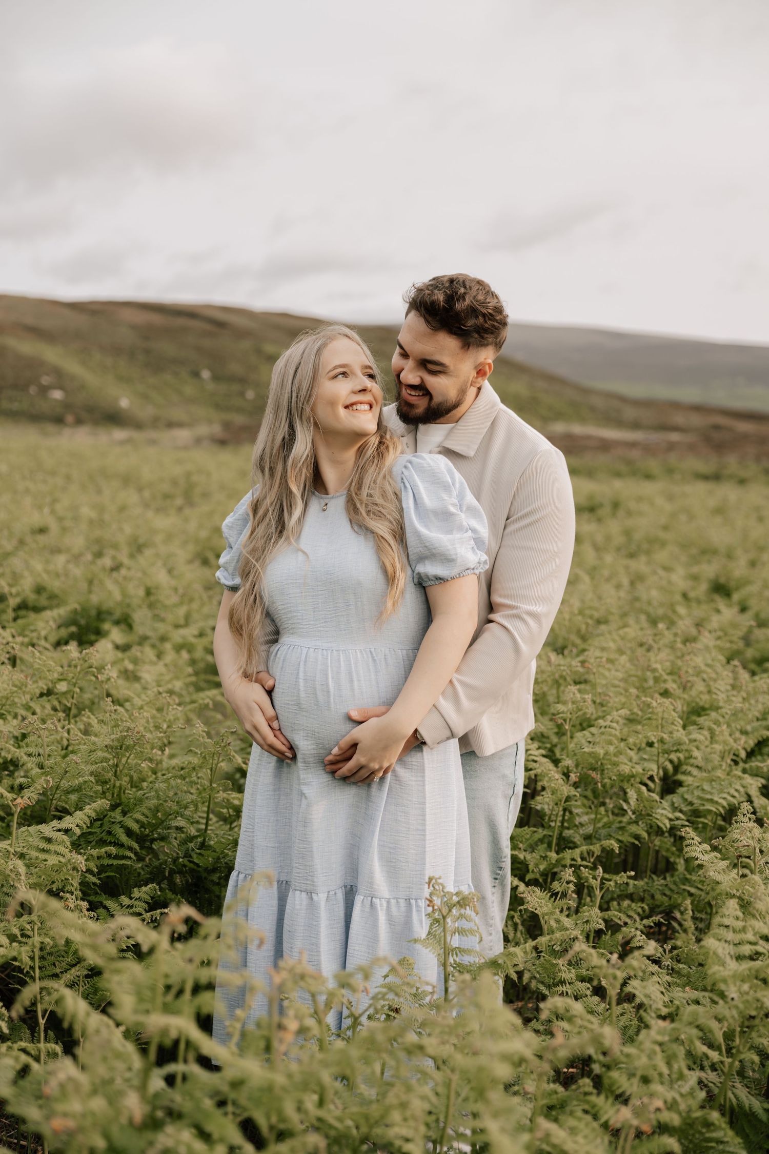 A couple shares a tender moment in a lush green field, the woman in a blue dress cradling her baby bump.