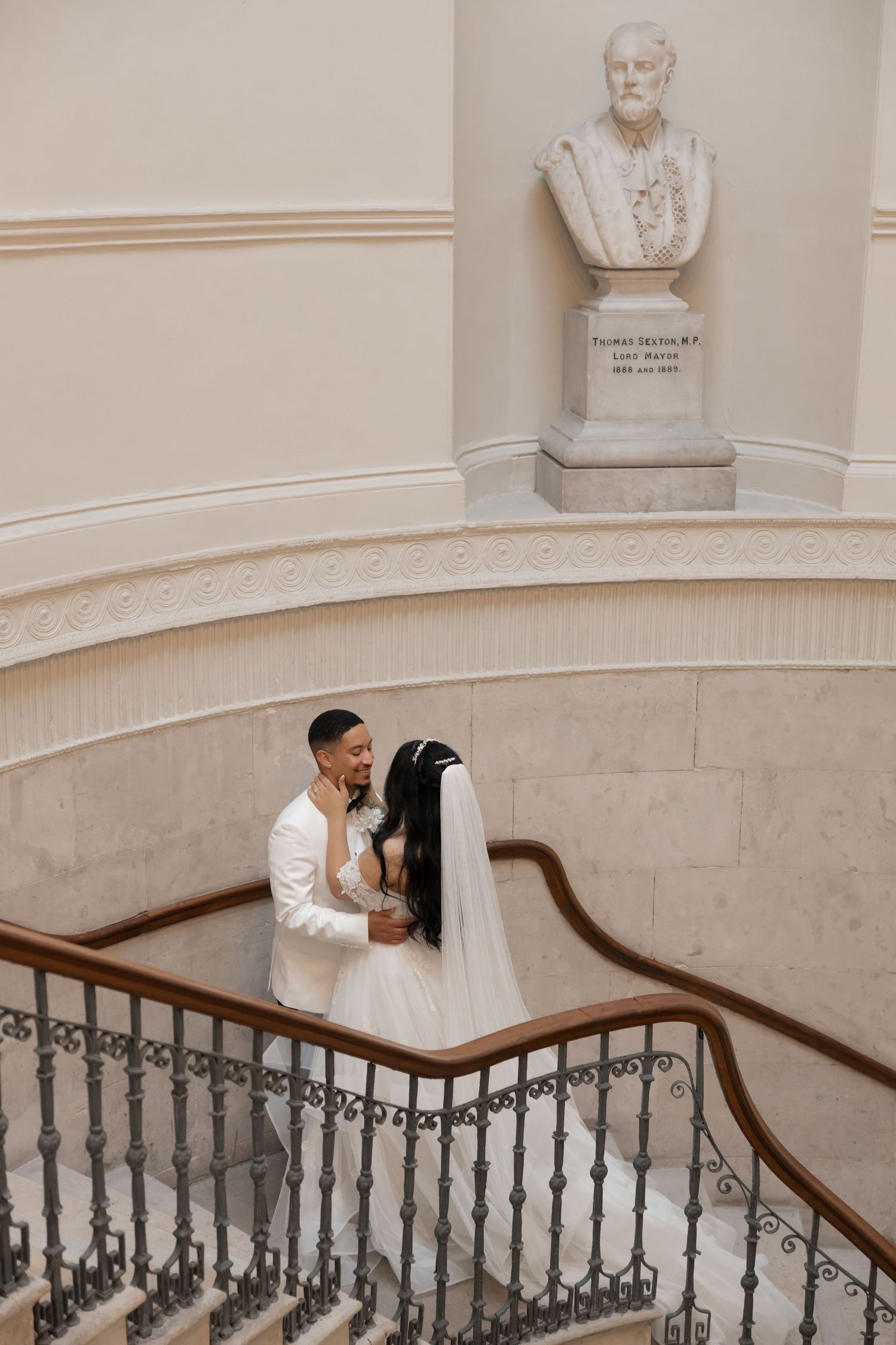 Elegant couple sharing an intimate moment on a grand staircase beneath a classical bust sculpture in a historic building.