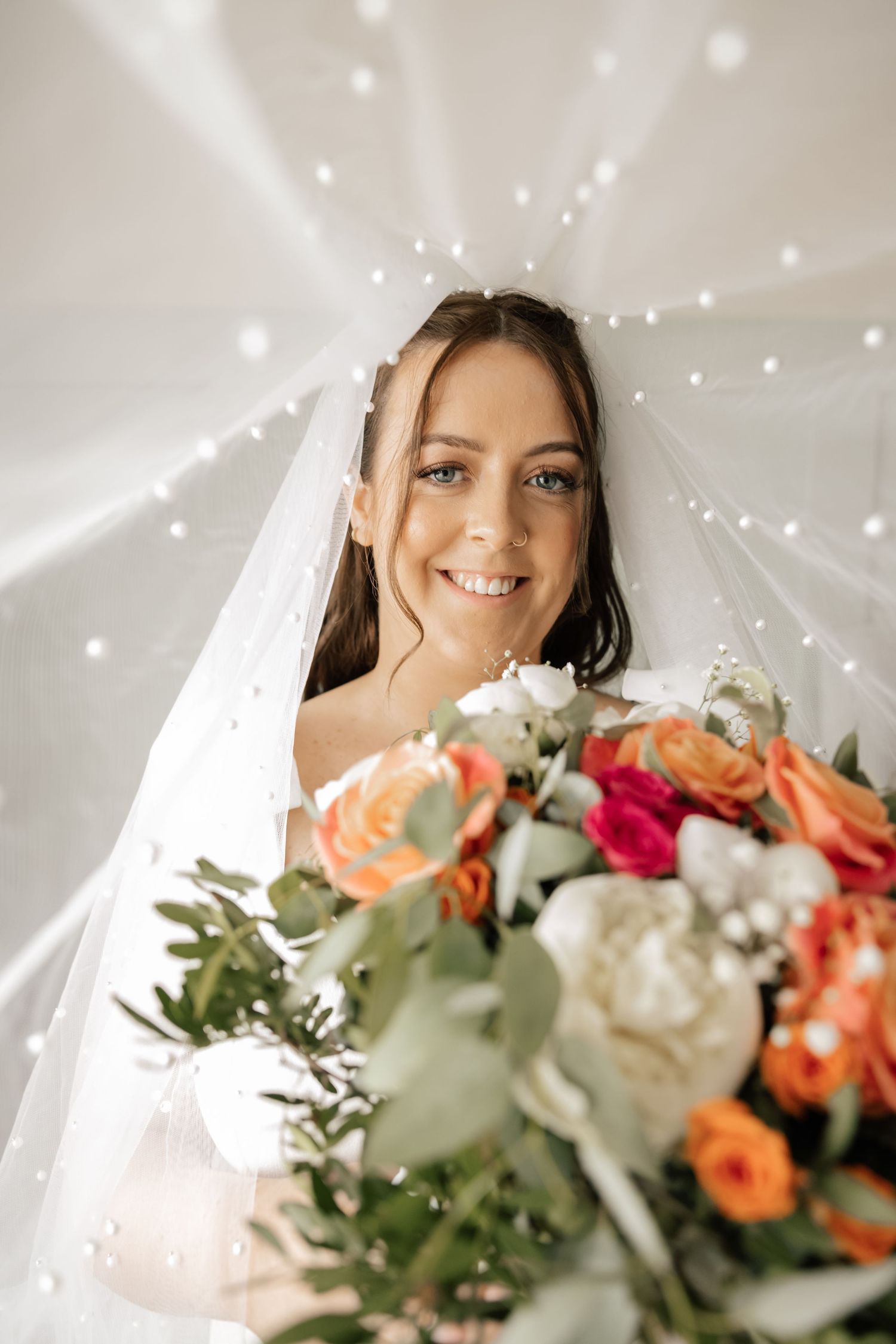 A bride in a white veil holds a vibrant bouquet of orange, red and white roses with greenery against a dreamy lit backdrop.