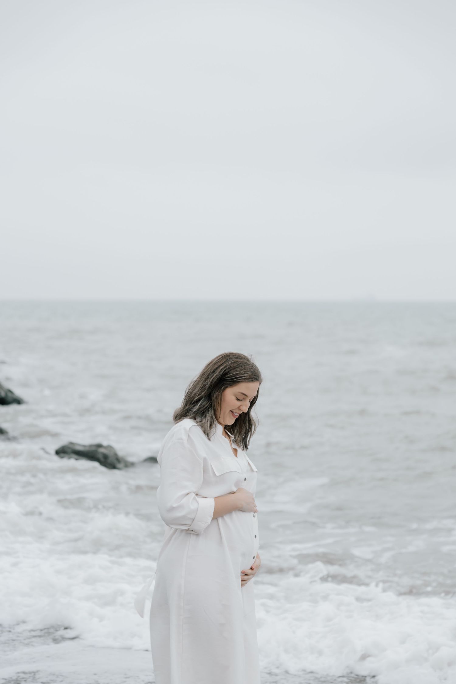 A pregnant woman in a white dress stands serenely by the ocean on a misty gray day.