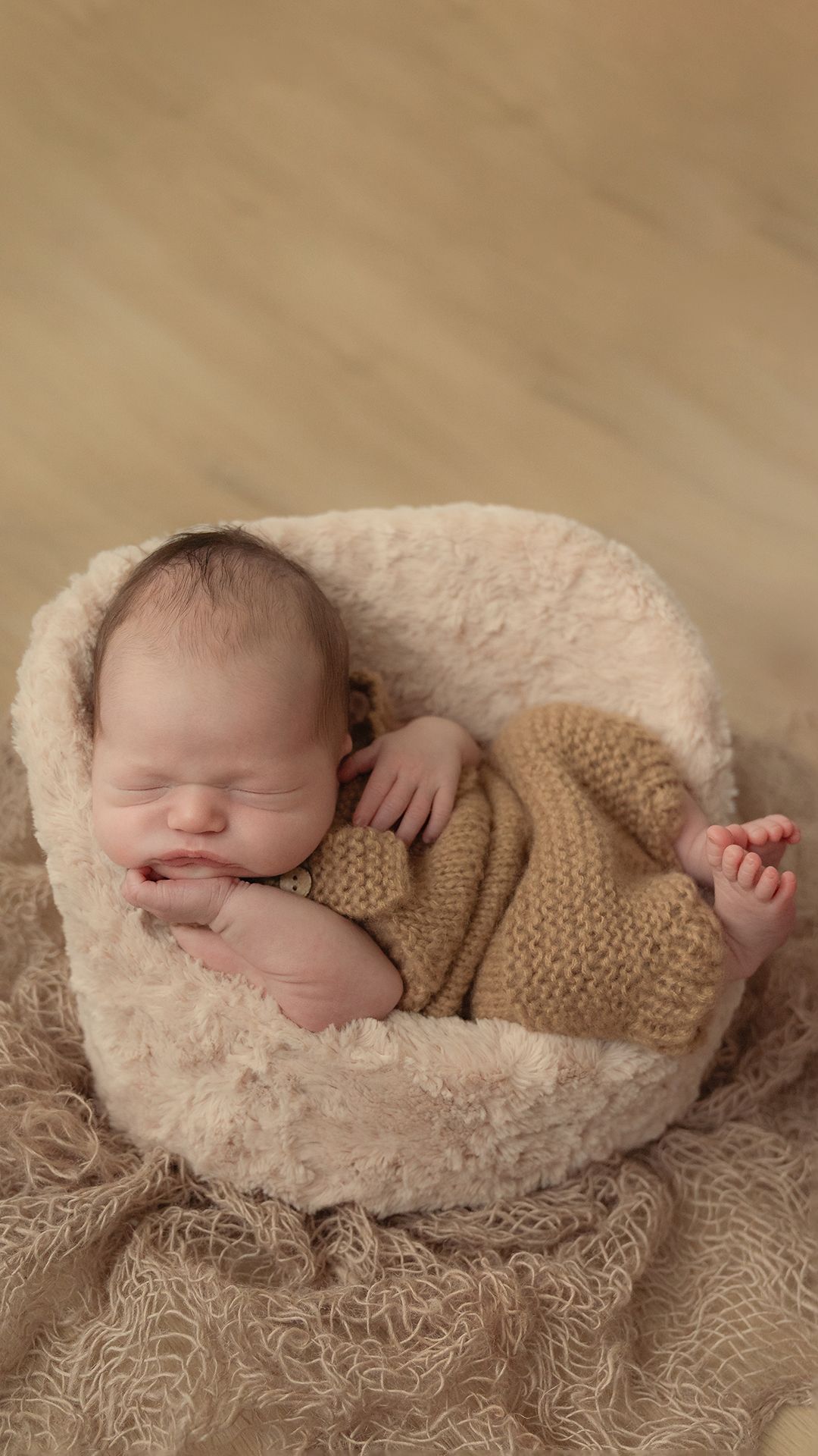 A sleeping newborn swaddled in a tan knit wrap rests peacefully on a cream-colored textured blanket.