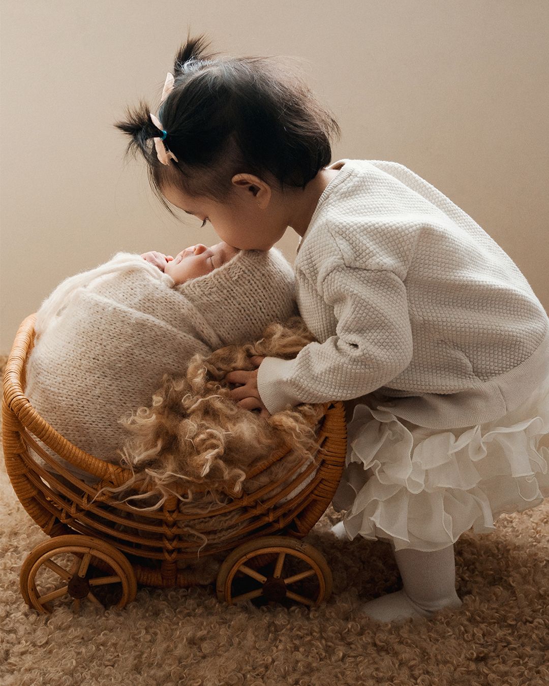 A toddler girl in a white dress gently kisses a newborn baby nestled in a wicker basket while newborn photographer in Vaughan taking picture