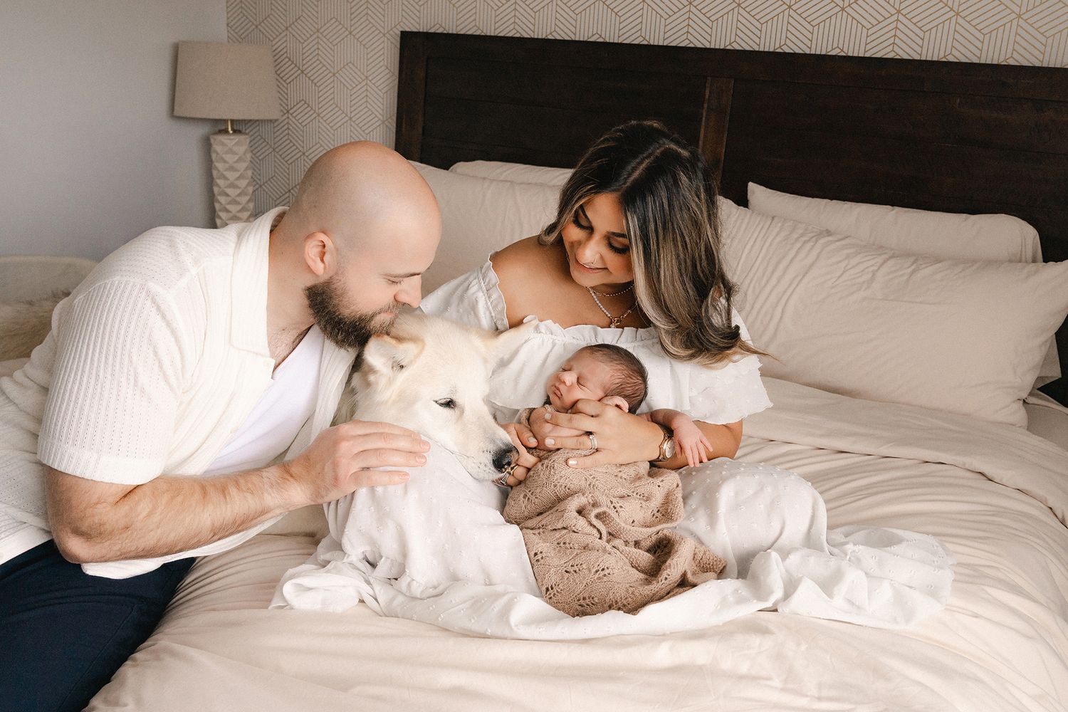 A couple sits on a white bed with their newborn baby and fluffy white dog in a cozy bedroom.