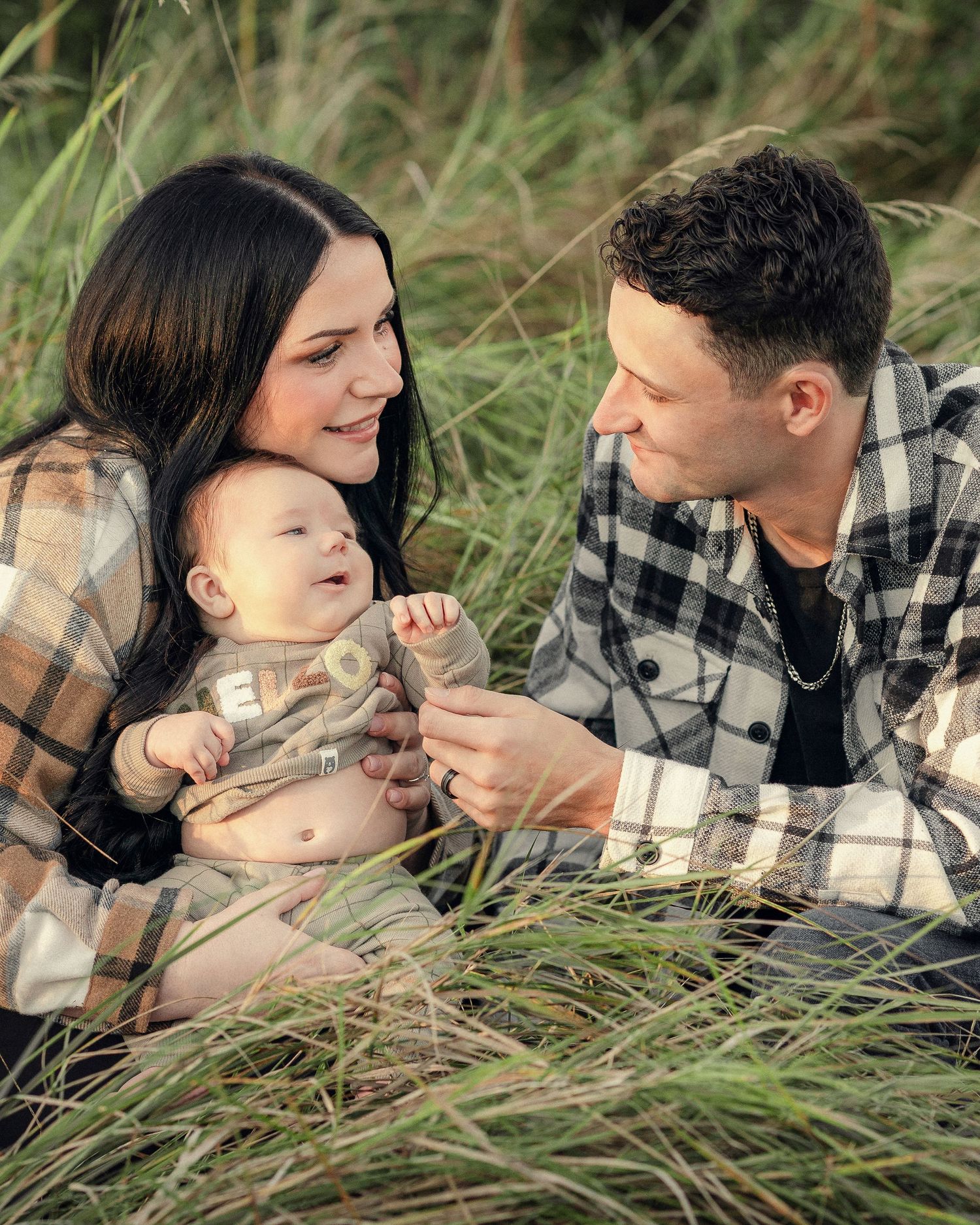 A couple lying in tall grass wearing matching plaid shirts share tender moments in a series of romantic photos.