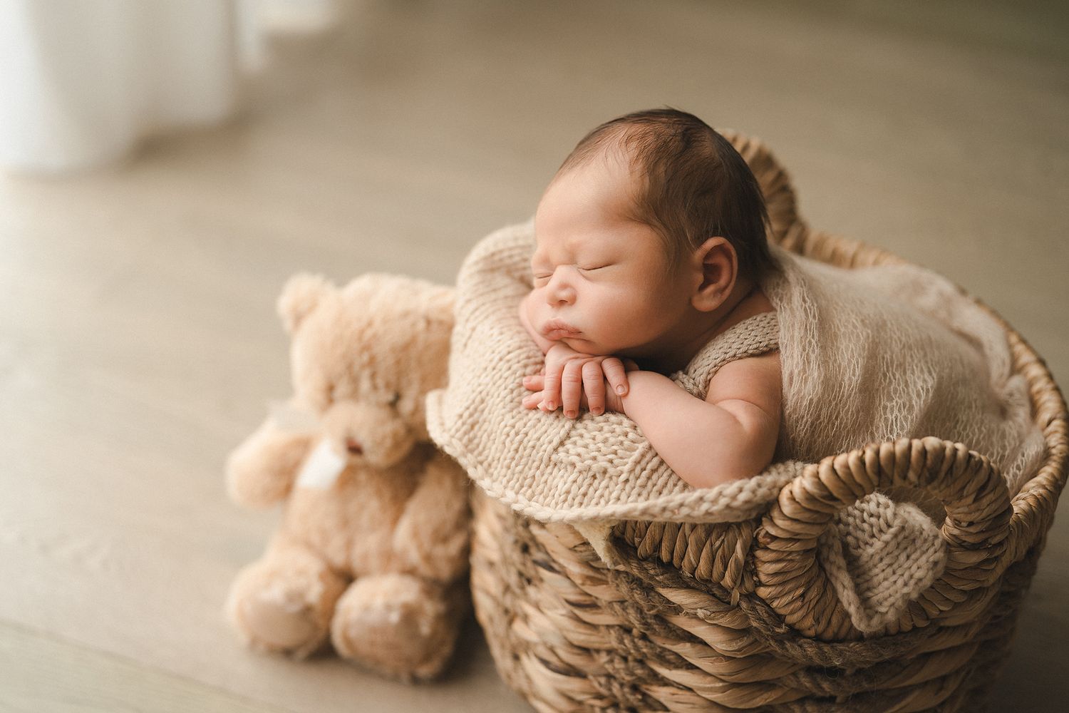 Sleeping newborn baby nestled in a wicker basket with a teddy bear, wrapped in a cozy beige knit blanket.