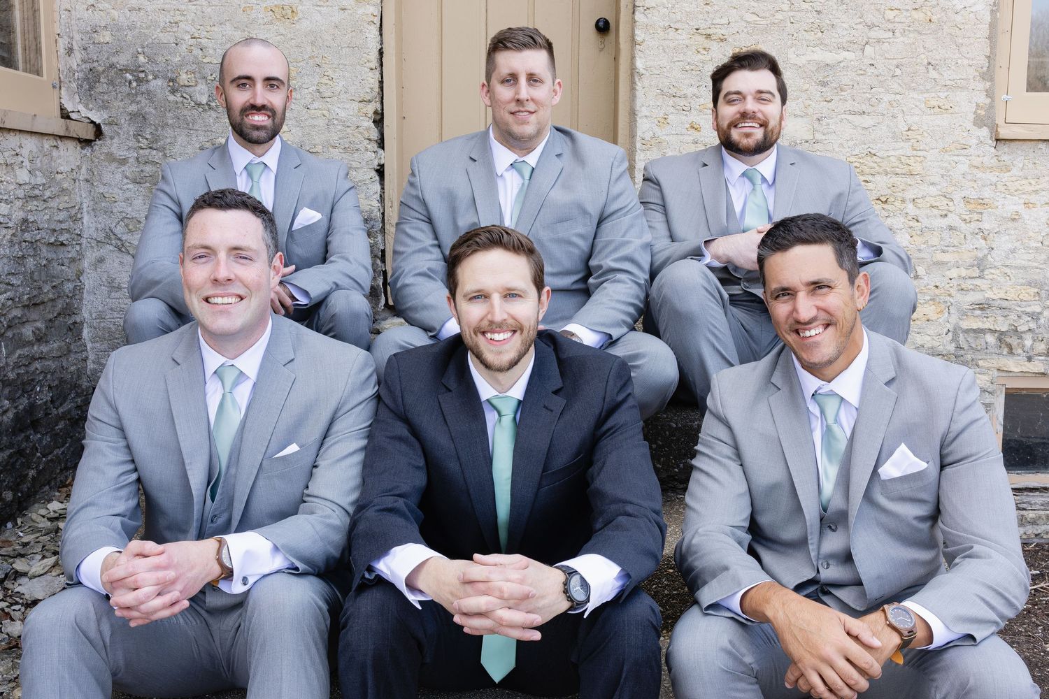 Six groomsmen in gray suits pose together outdoors, with the groom in a navy suit and green tie in the center.