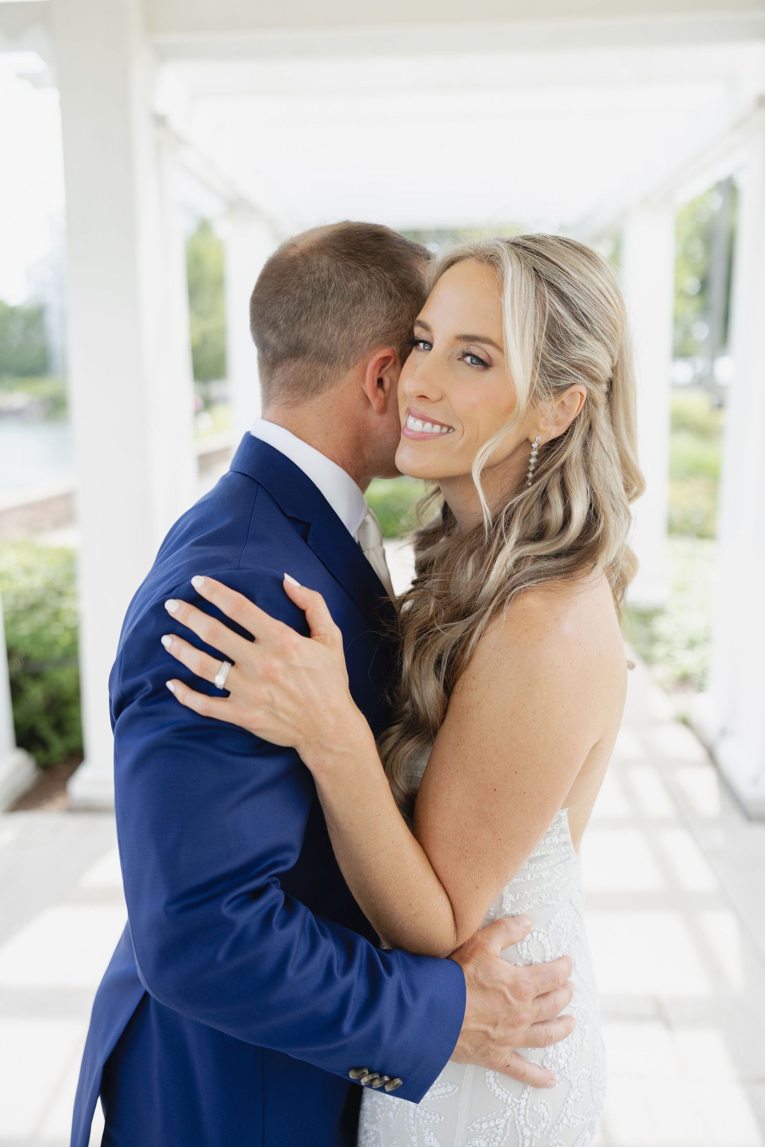 Bride in white gown smiles over shoulder as groom in blue suit embraces her under white columns.